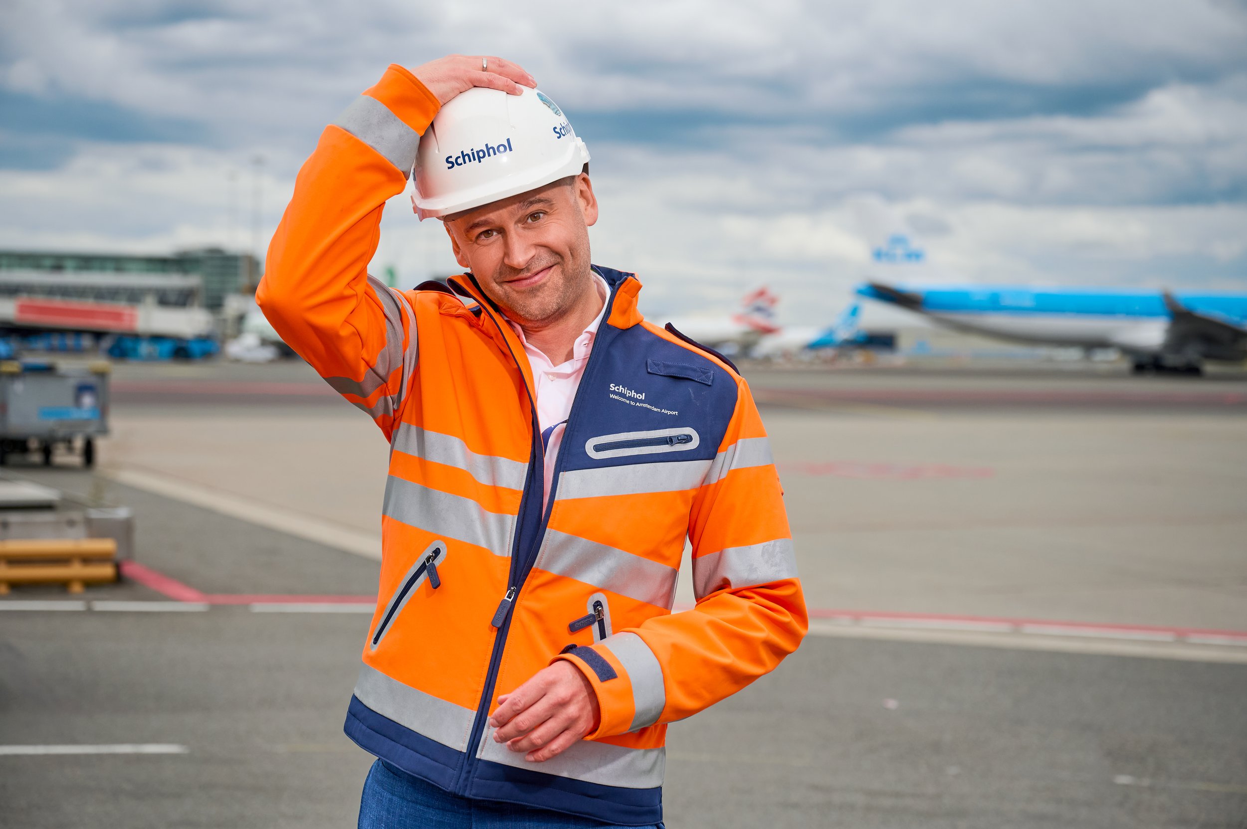 Man in oranje veiligheidshes met cap met 'Schiphol' op, op luchthaven naast vliegtuigen.