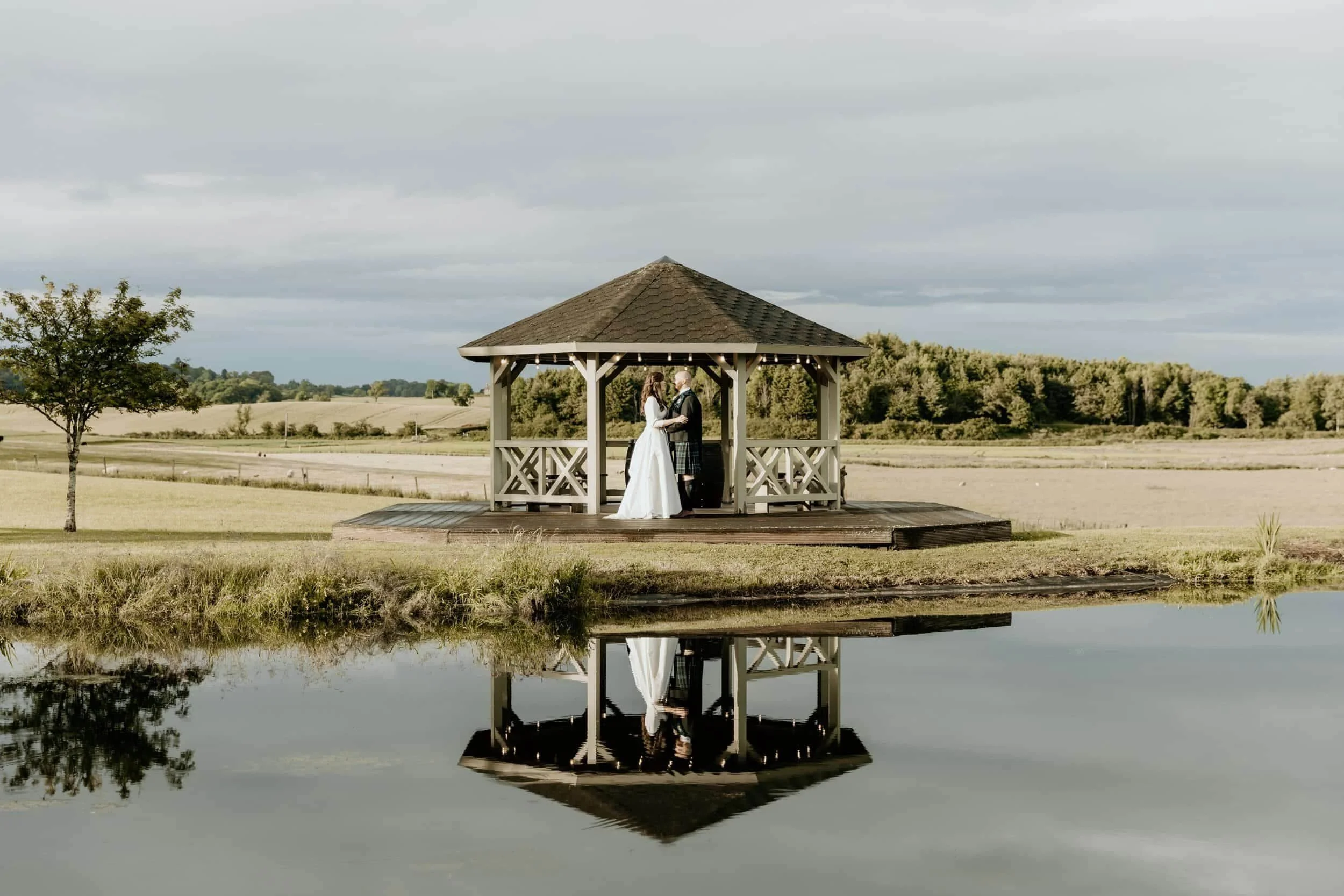 A bride and groom standing in a gazebo by a pond, holding hands and looking at each other, with a open countryside landscape in the background.