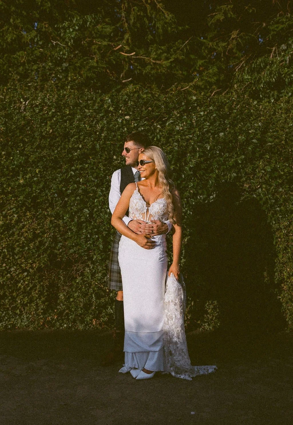 A bride and groom standing close together outdoors, smiling, with a green hedge in the background. They are wearing sunglasses and the groom is dressed in traditional Scottish attire.