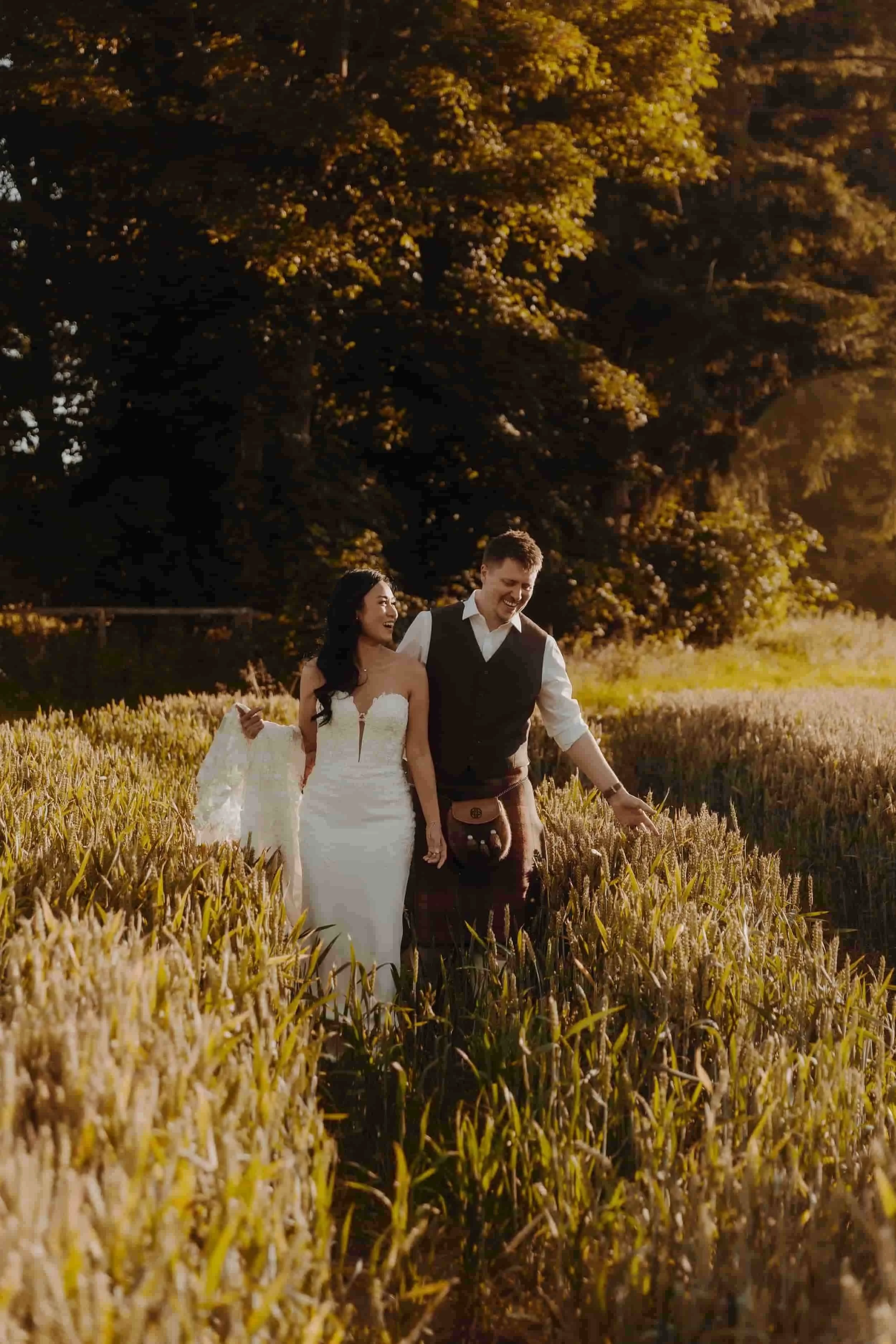 A couple in wedding attire walking through a field of tall grass or wheat, holding hands and smiling, with large trees and sunset light in the background.
