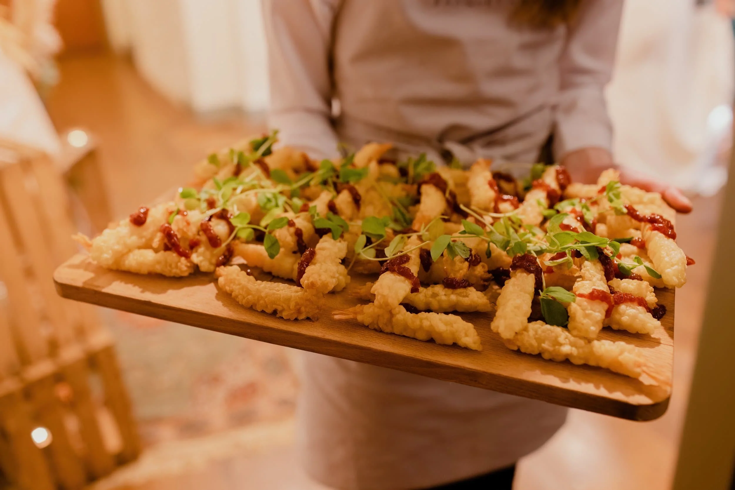 A person holds a wooden tray with crispy appetizers topped with greens and sauce, perfect for catering and ready to be served.