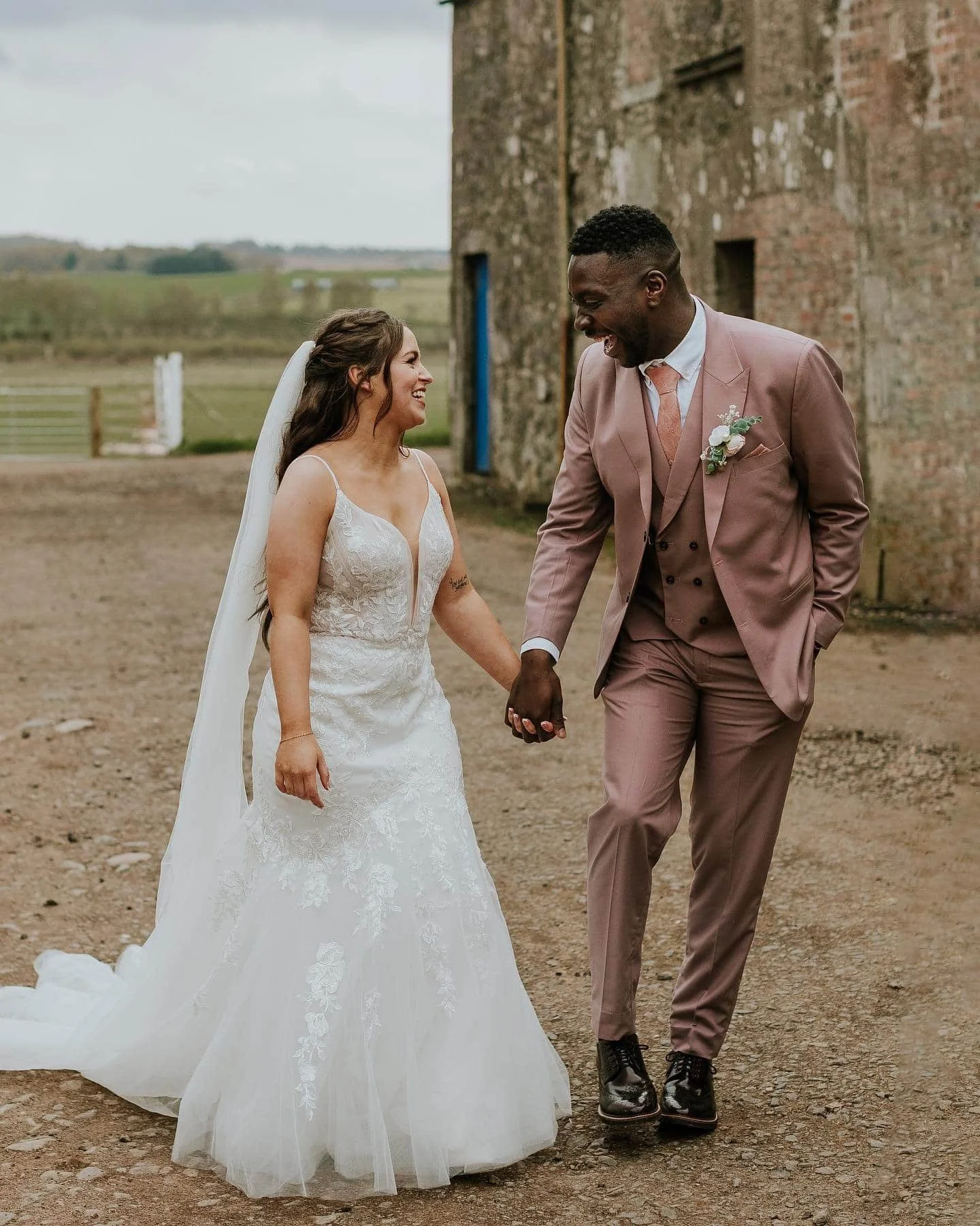 A bride and groom holding hands and smiling at each other outdoors with a rustic brick building in the background.