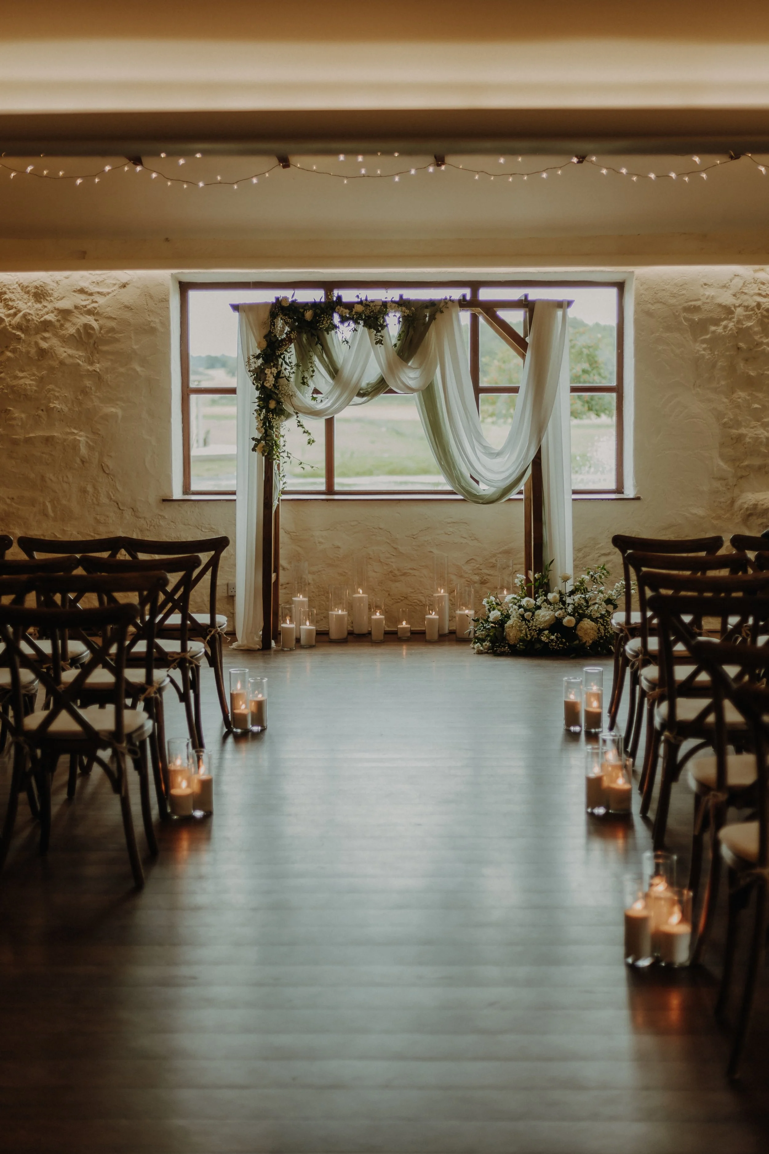 Interior of a wedding ceremony setup with rows of wooden chairs, a decorated arch with white fabric and flowers, and candles on the floor, in front of a window overlooking a green landscape.