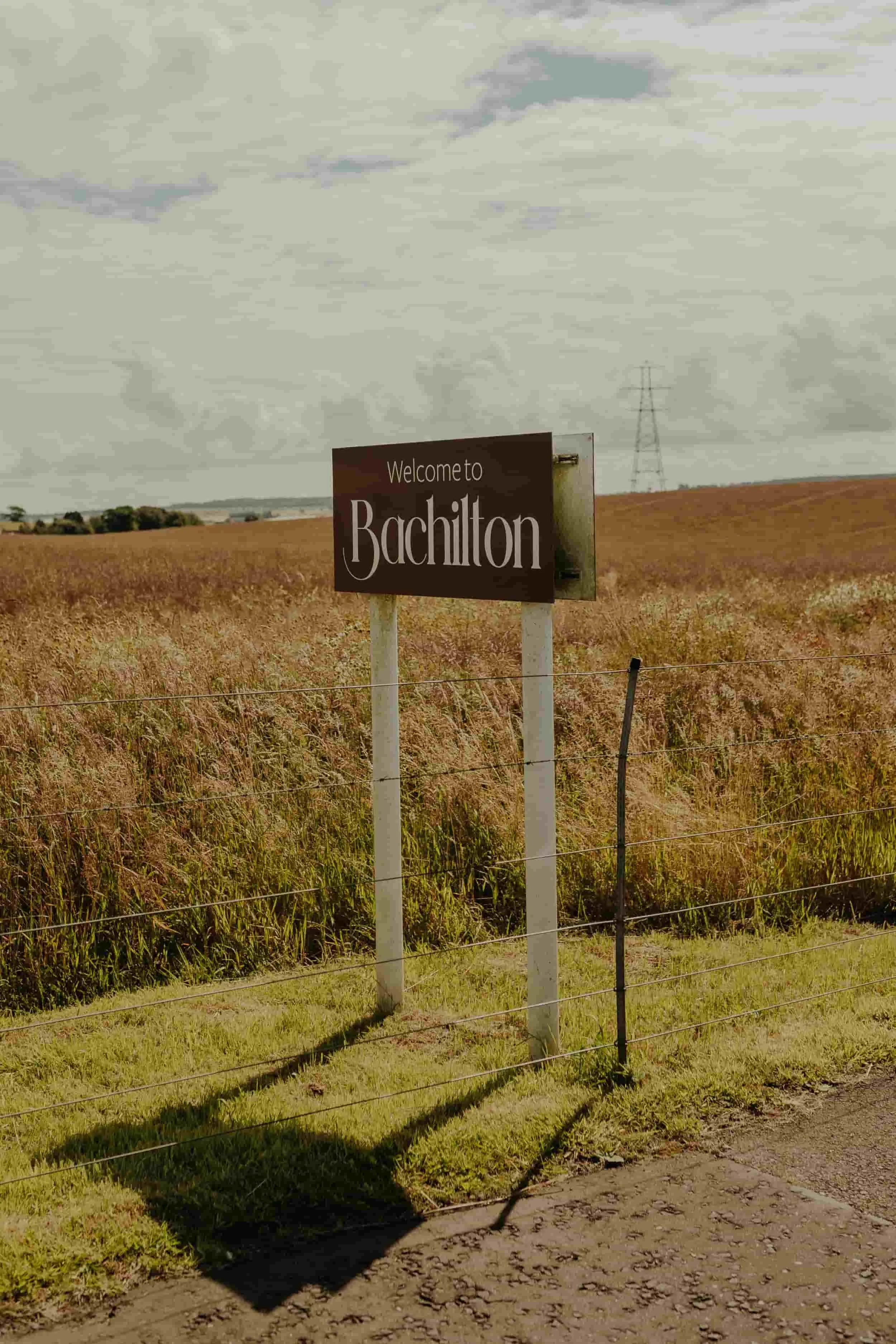 A sign reading Welcome to Bachilton stands by a grassy field under a cloudy sky.