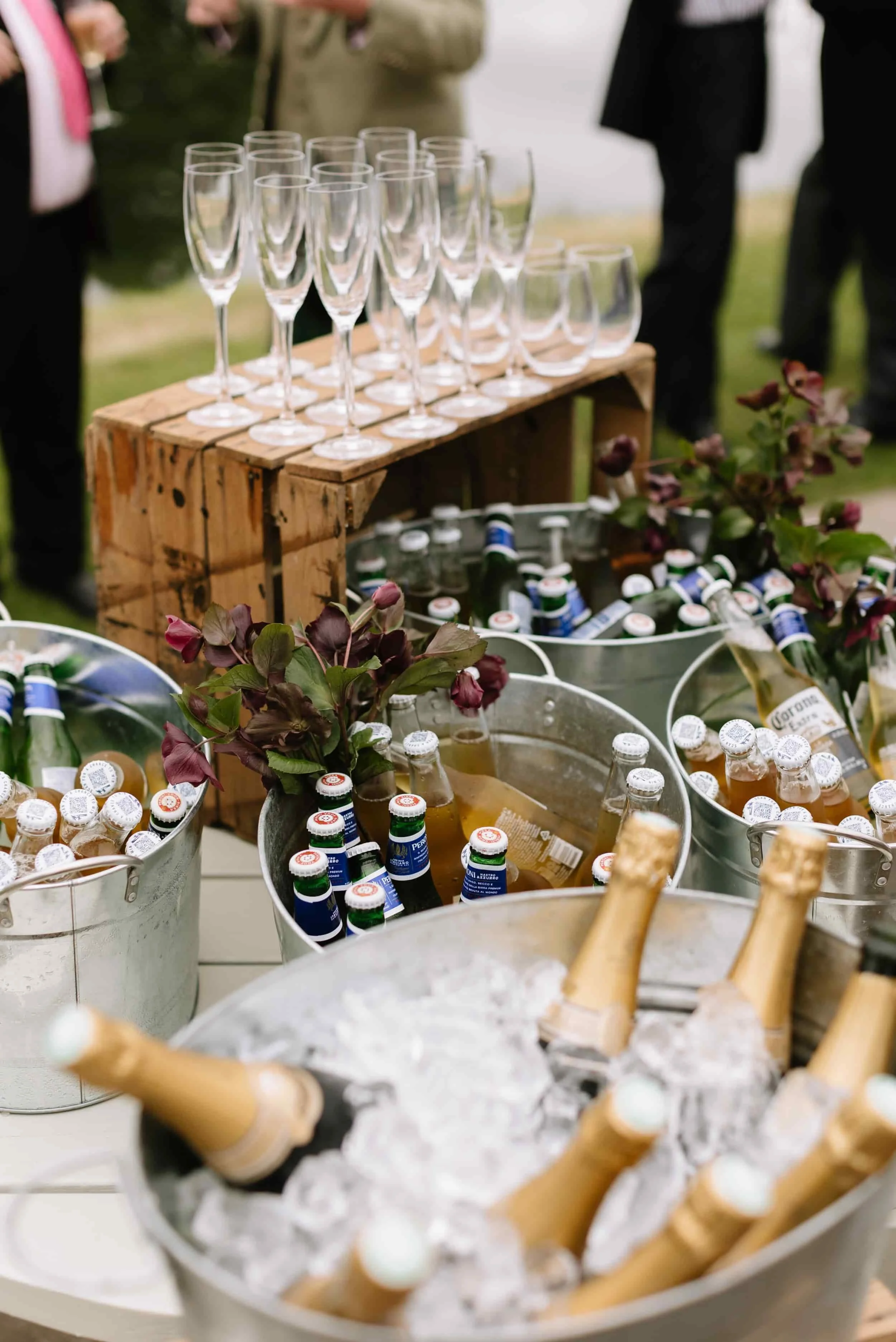 Champagne and beer bottles in ice buckets, empty champagne glasses on a wooden crate, and people in formal attire at an outdoor event.