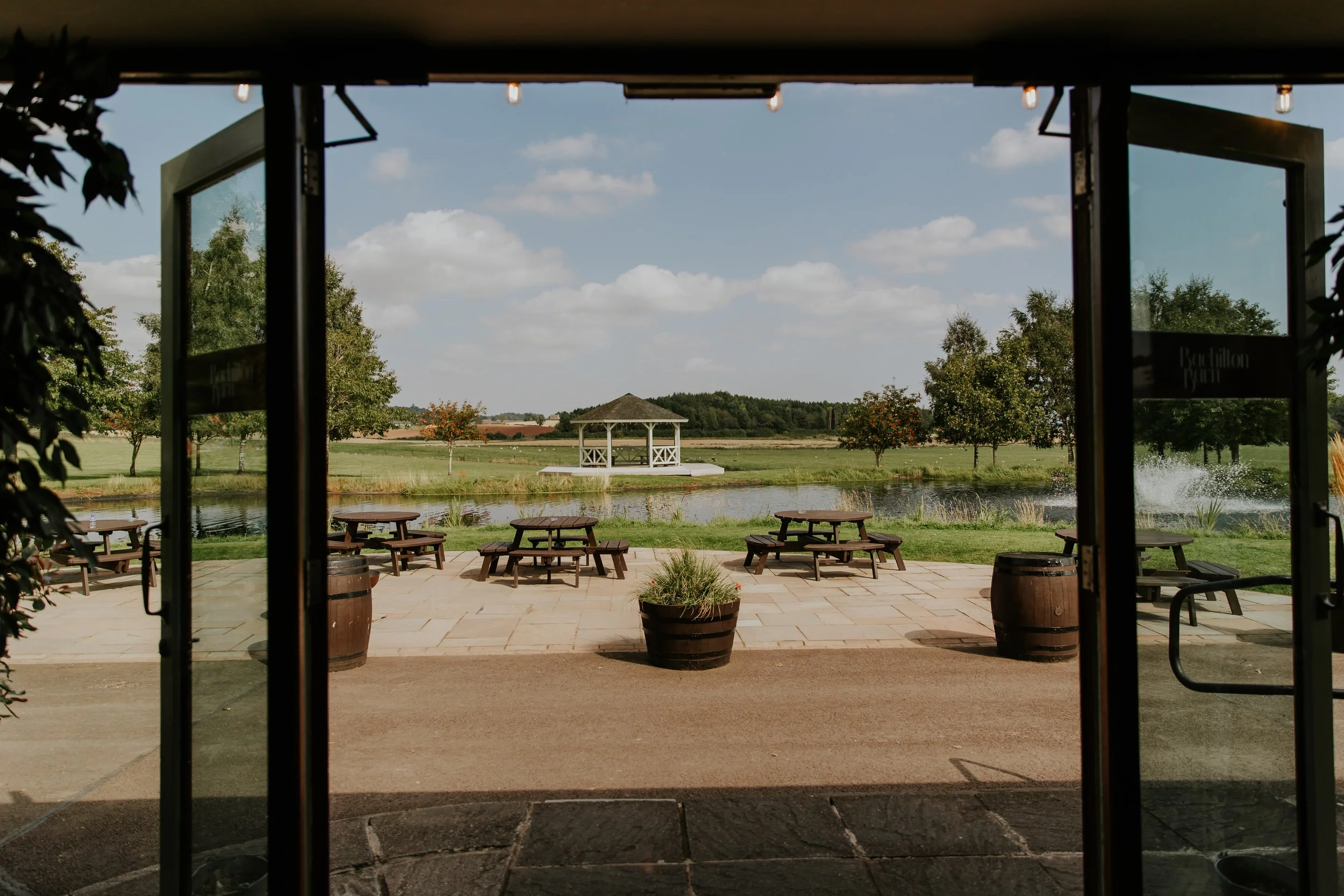 View through open doors to an outdoor patio with picnic tables, a pond, trees, and a gazebo in the distance under a partly cloudy sky.