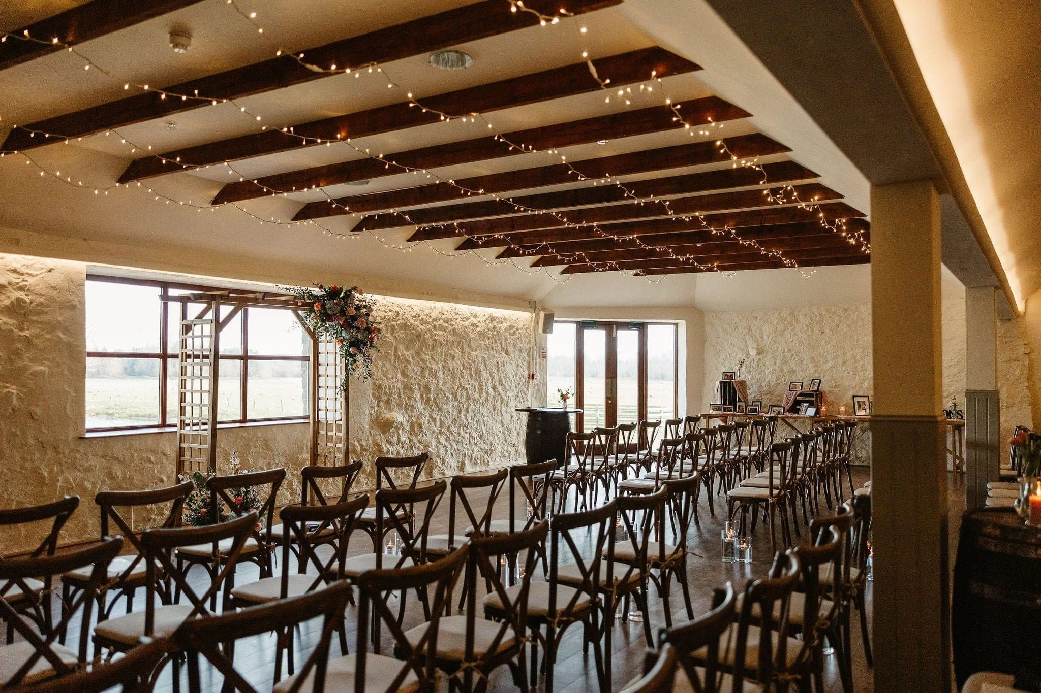 A rustic wedding ceremony room with wooden chairs, string lights, and floral decorations, set up facing large windows.