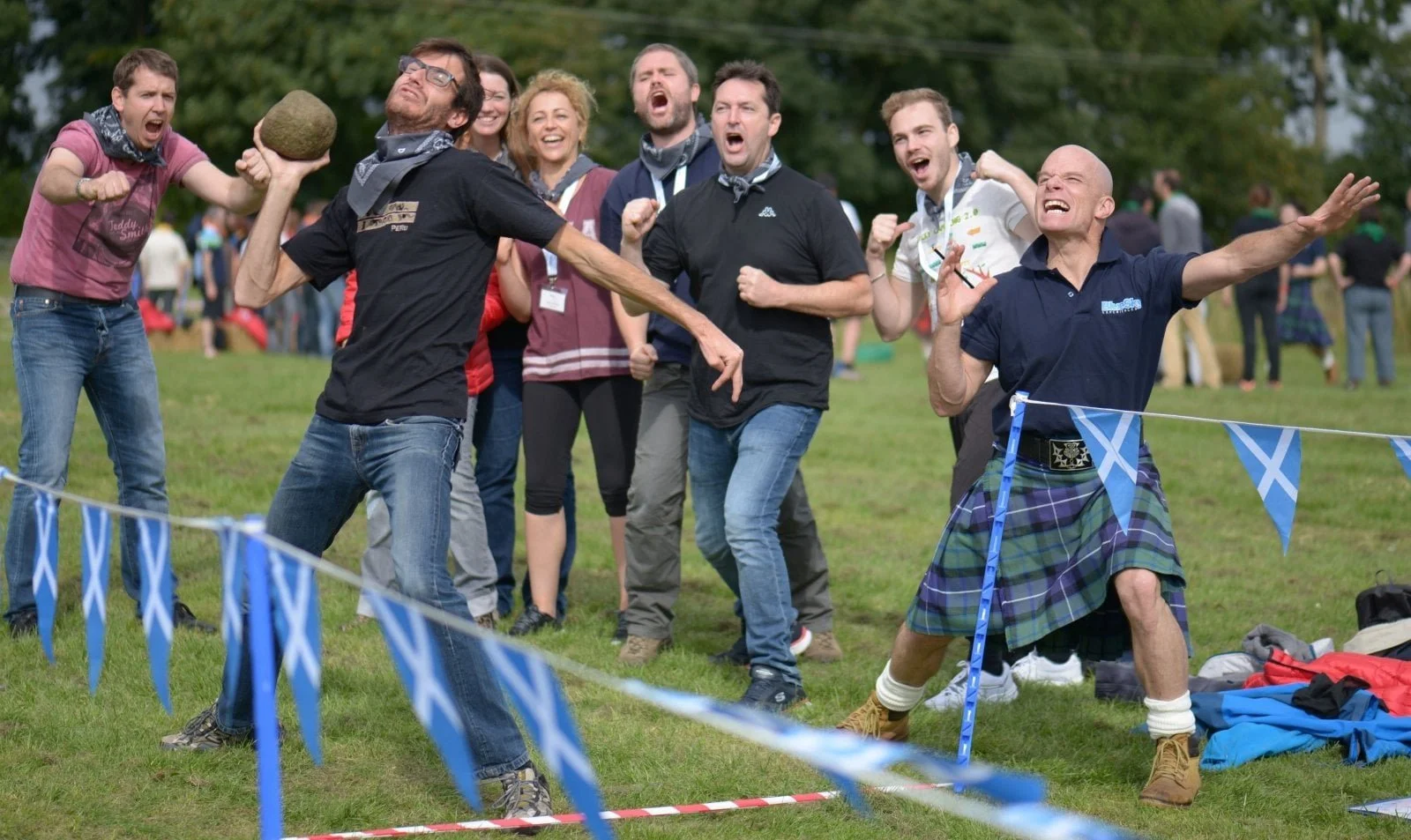 A man in a kilt and another man throw stones as a cheering crowd watches at an outdoor event with blue and white flags.