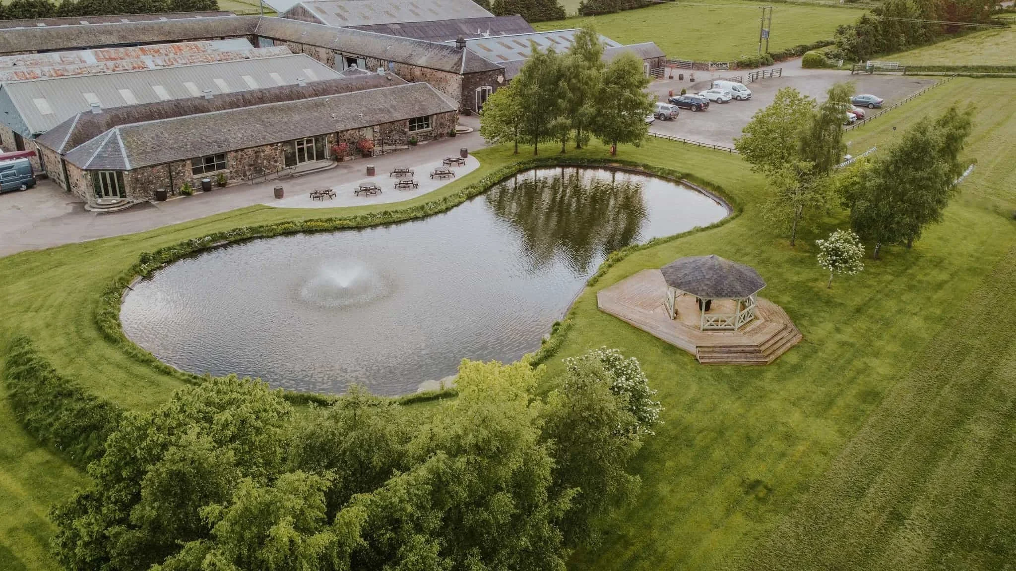 Aerial view of a property with a pond featuring a water fountain, a gazebo on a wooden deck, surrounded by trees, a stone building with a patio, and a parking lot with parked cars.