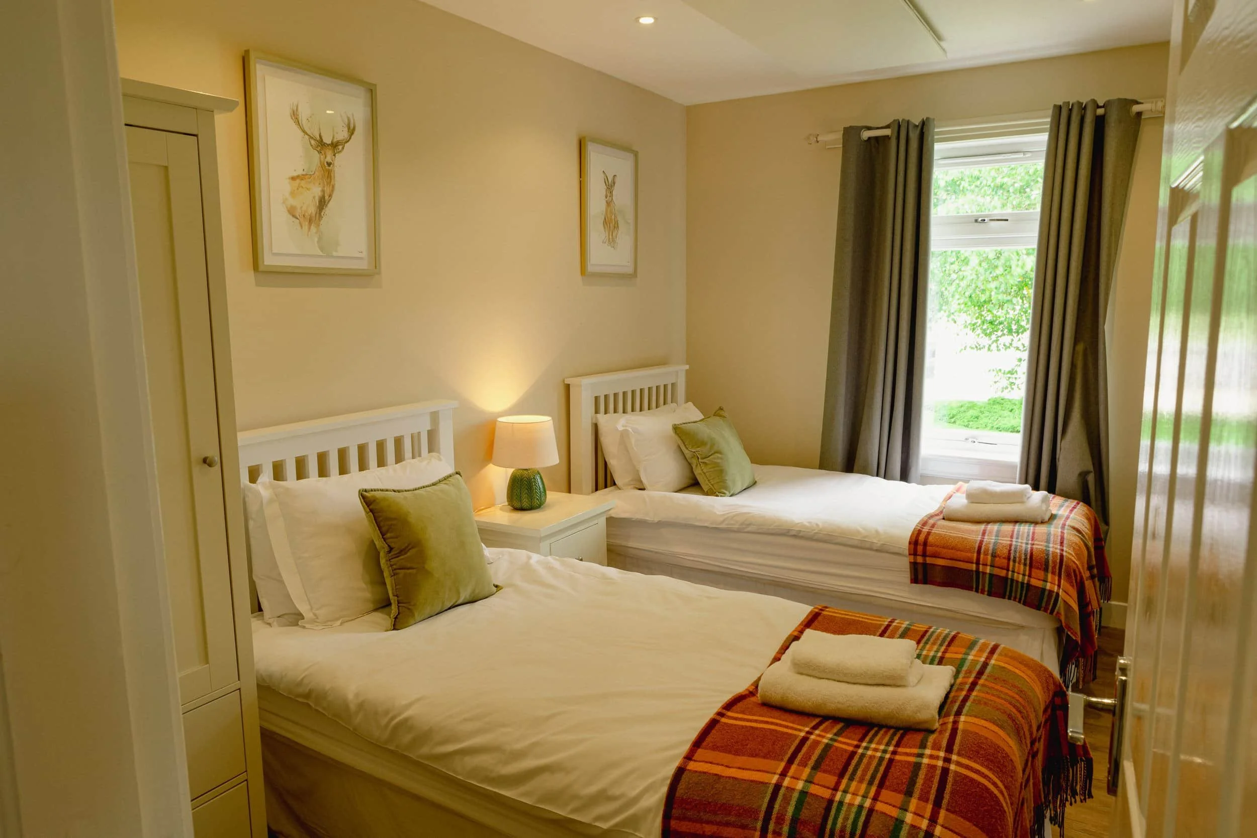 A bedroom with two single beds, each topped with white linens and green cushions. There's a bedside table with a lamp, framed artwork of deer on the wall, a window with gray curtains, and folded towels with plaid blankets at the foot of each bed.