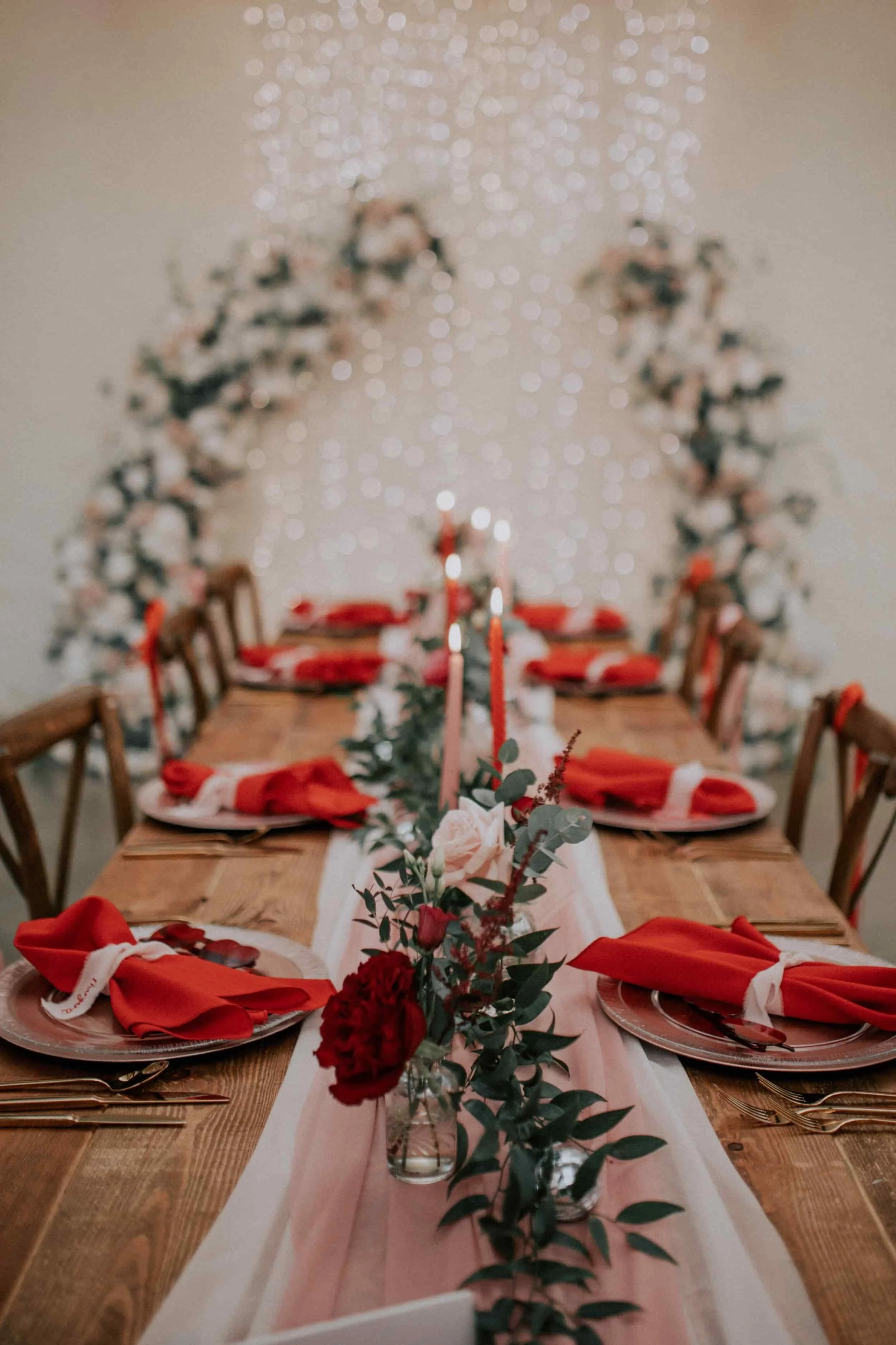 A beautifully decorated dinner table with red napkins, gold flatware, pink candles, and floral centerpieces, set for a festive event. In the background, there is a floral arch and string lights creating a warm ambiance.