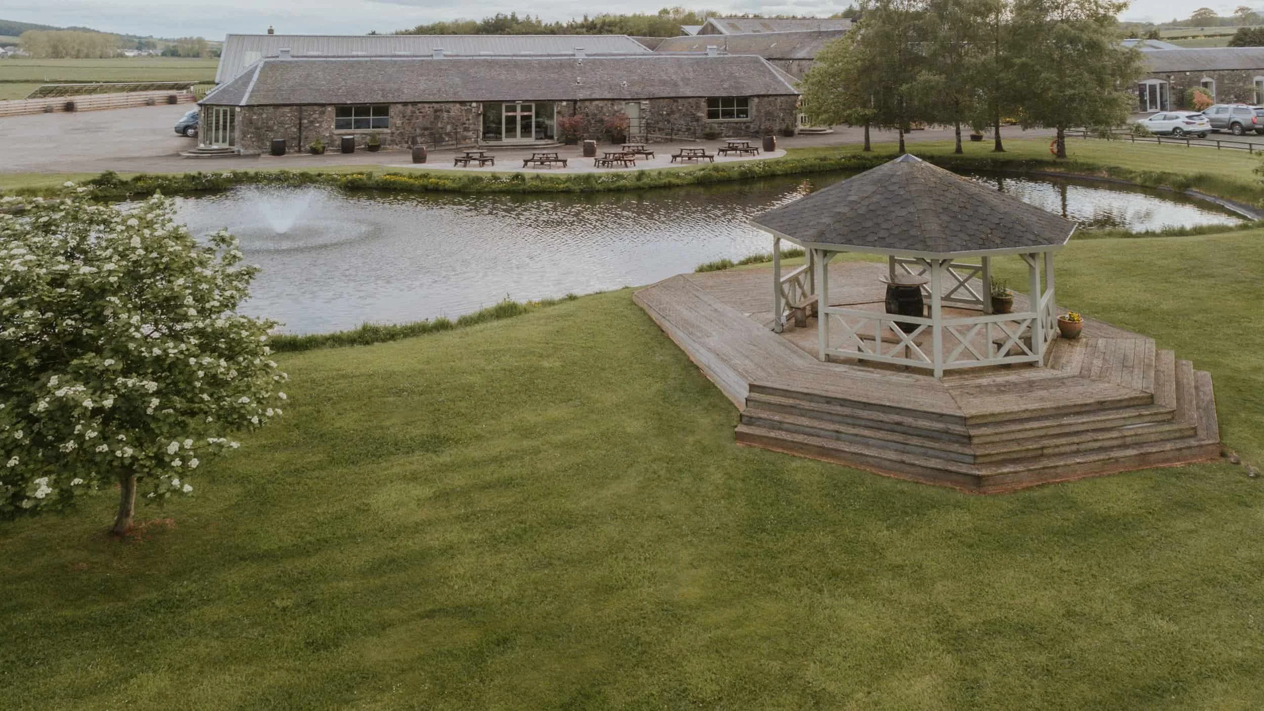 A wooden gazebo stands by a small pond with a fountain, near a stone building and picnic tables on a grassy lawn.