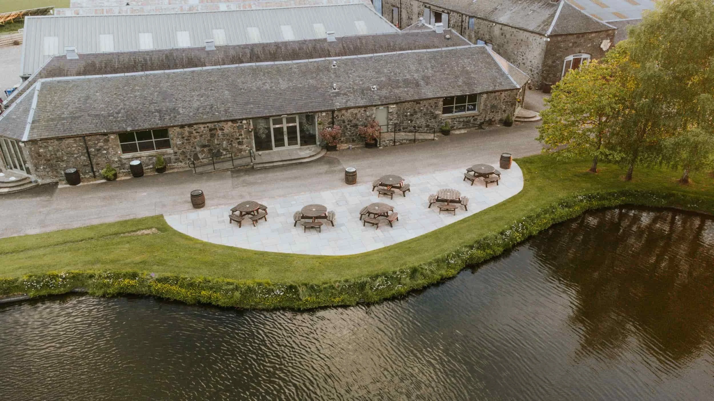 A stone building with a metal roof, several windows, and a glass door, is situated near a body of water. There are outdoor picnic tables, trash bins, and potted plants on a paved and grassy area close to the building, with trees on the side.