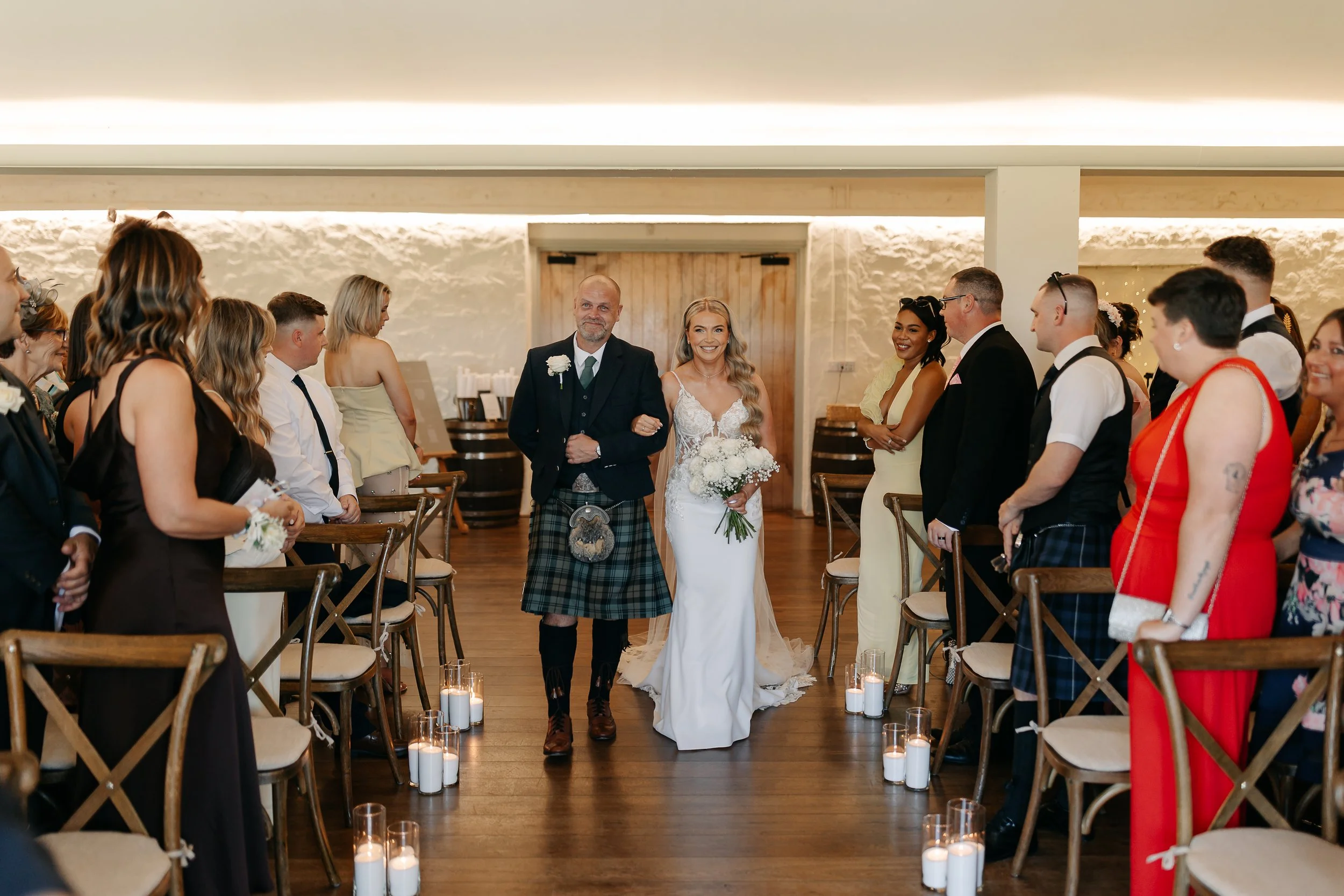 A bride walking down the aisle escorted by a man in traditional Scottish attire at a wedding ceremony. Guests are standing on both sides, watching and smiling, with lit candles along the aisle.