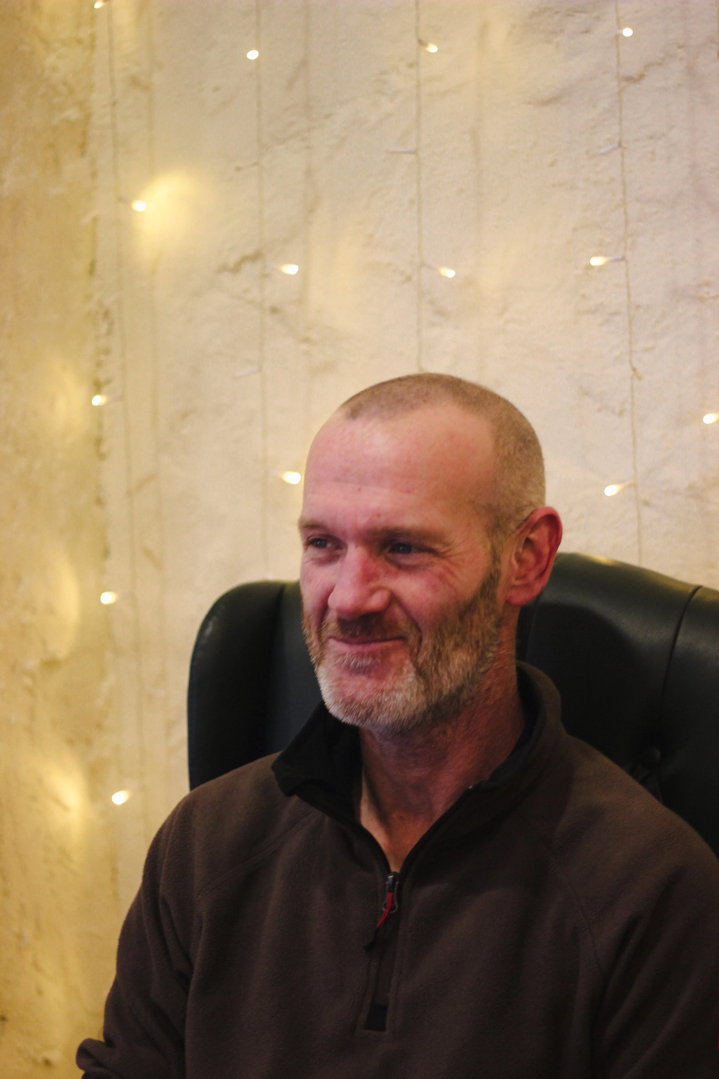 A middle-aged man with a beard sitting in a black chair, smiling softly, against a background of beige textured wall with small, warm, white string lights.