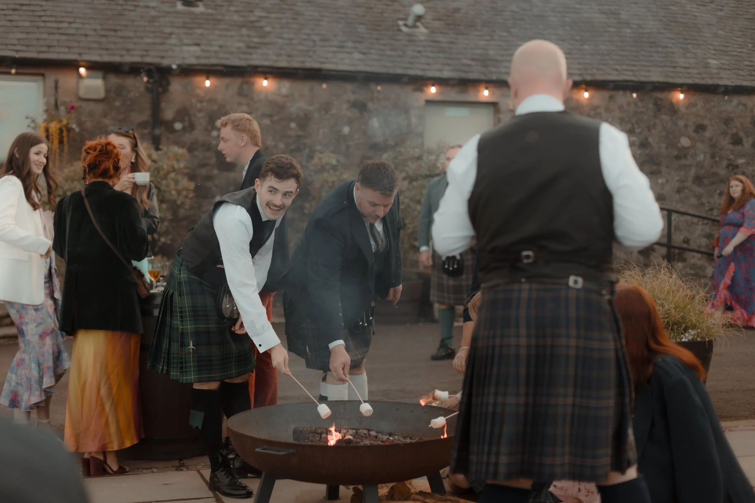 People gathered around a fire pit roasting marshmallows at an outdoor celebration, some dressed in kilts.