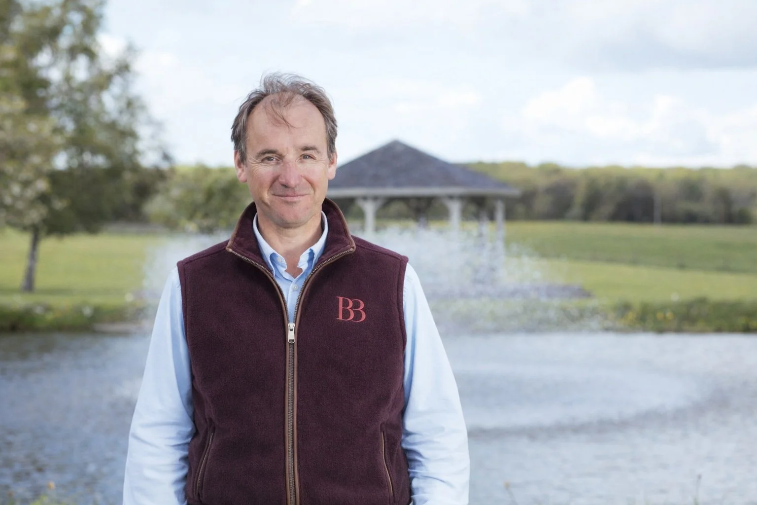 A man stands outdoors near a body of water with a pavilion in the background, wearing a maroon vest with a logo, a light blue shirt, and smiling at the camera.