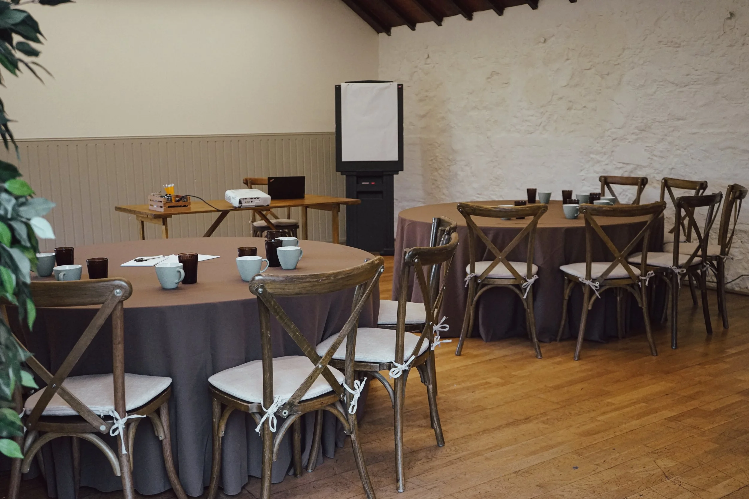 A room set up for a meeting or event with round tables covered in brown tablecloths, surrounded by wooden chairs with white cushions and tied ribbons. There is a projector on one of the tables, a flip chart on a stand, and cups and glasses on the tables. The room has wooden floors, a textured white wall, and a ceiling with exposed wooden beams.
