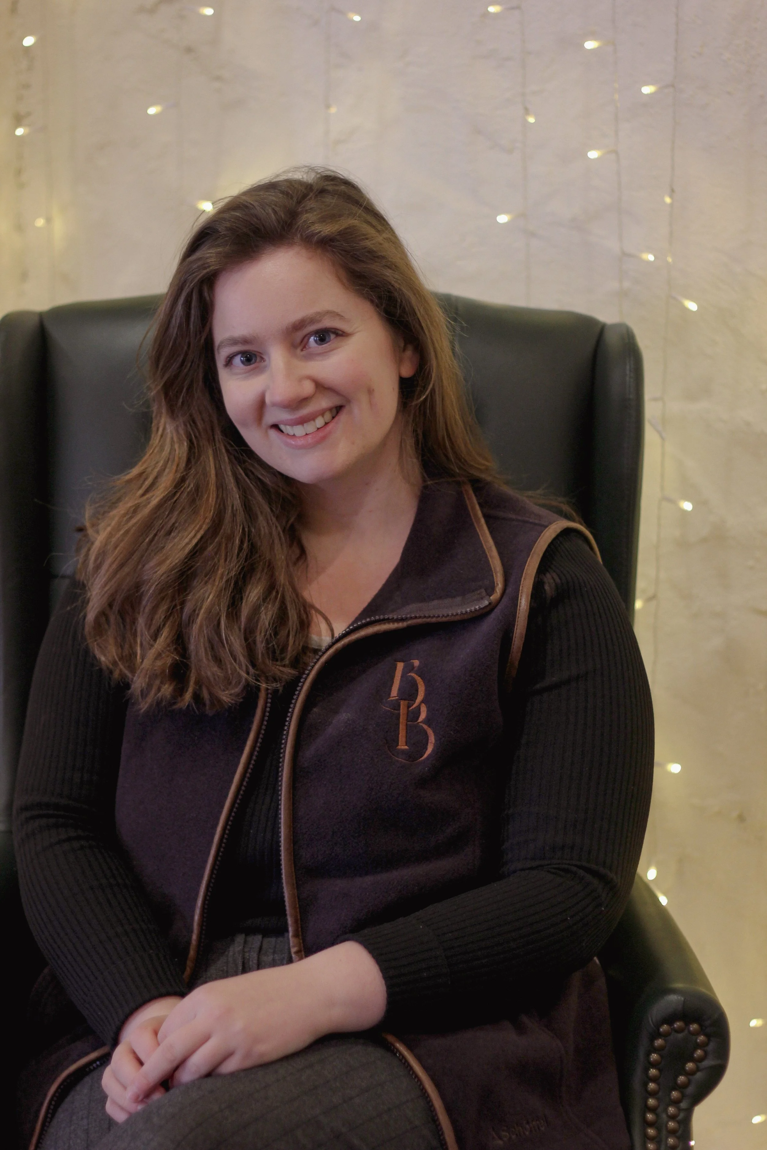 A woman with long brown hair smiling, sitting in a black leather armchair, in front of a white wall with string lights.