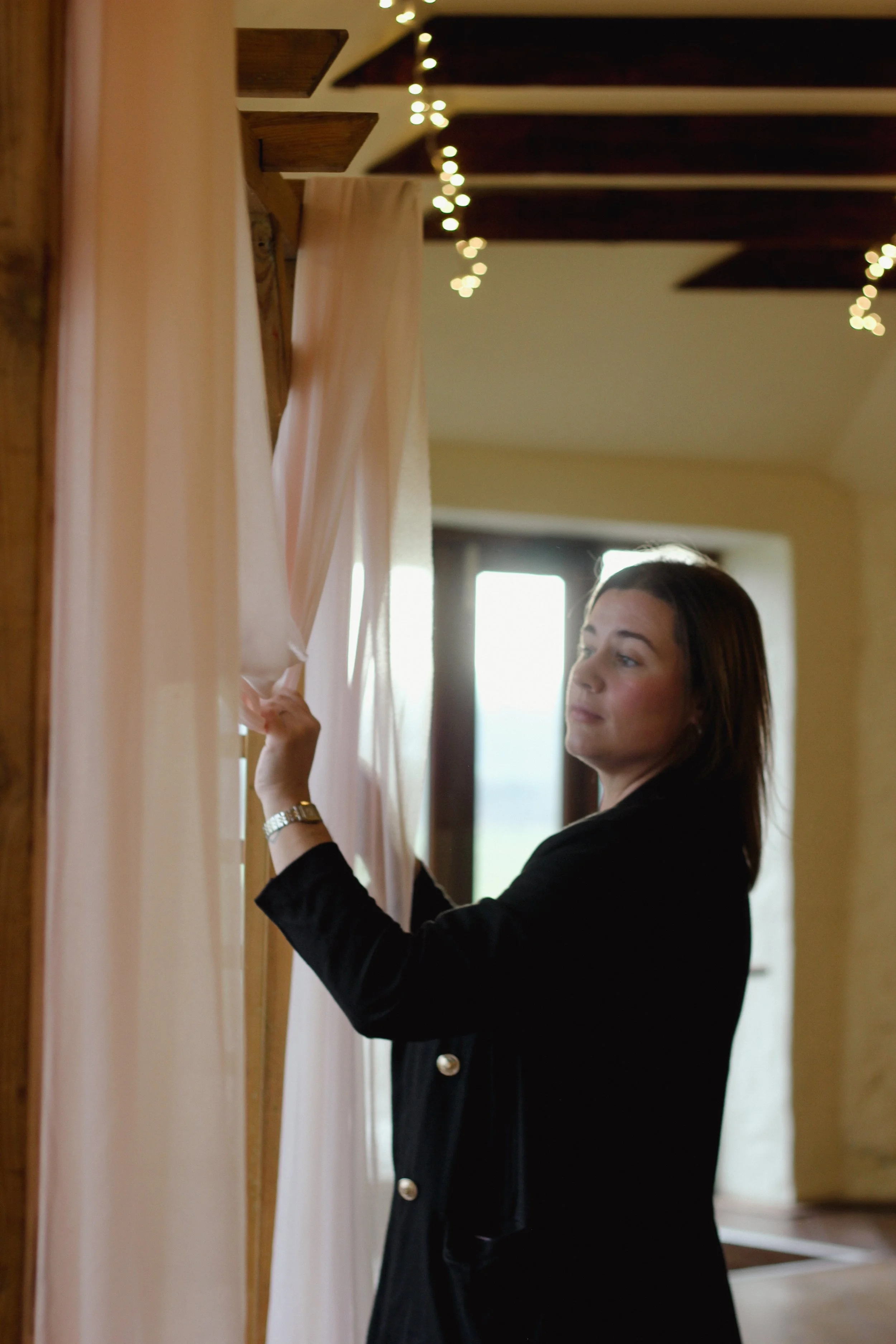 Woman in black jacket adjusting light pink curtains in a room with large windows and string lights on the ceiling.