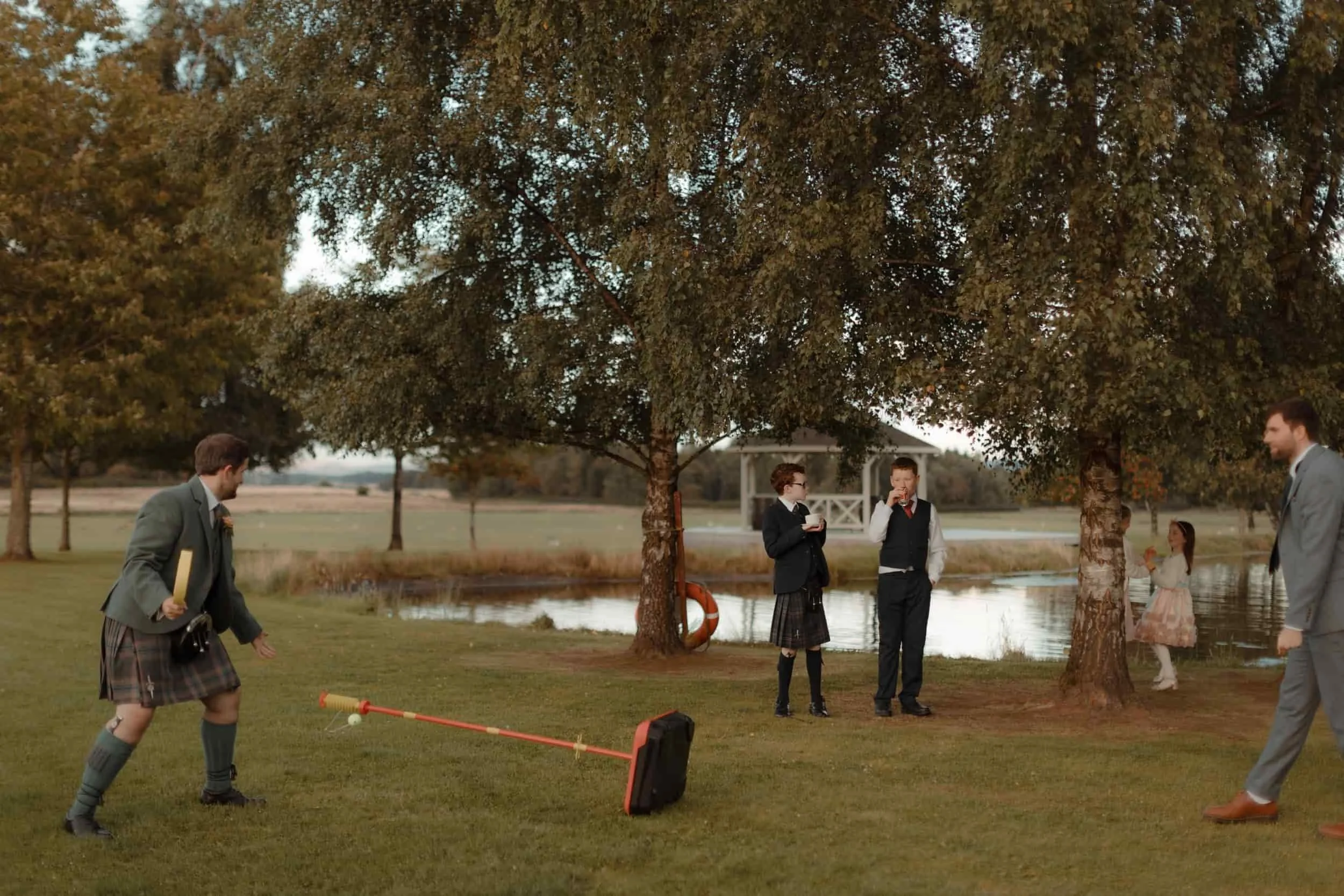 Group of children and adults playing outside near a pond with trees in the background during daytime.