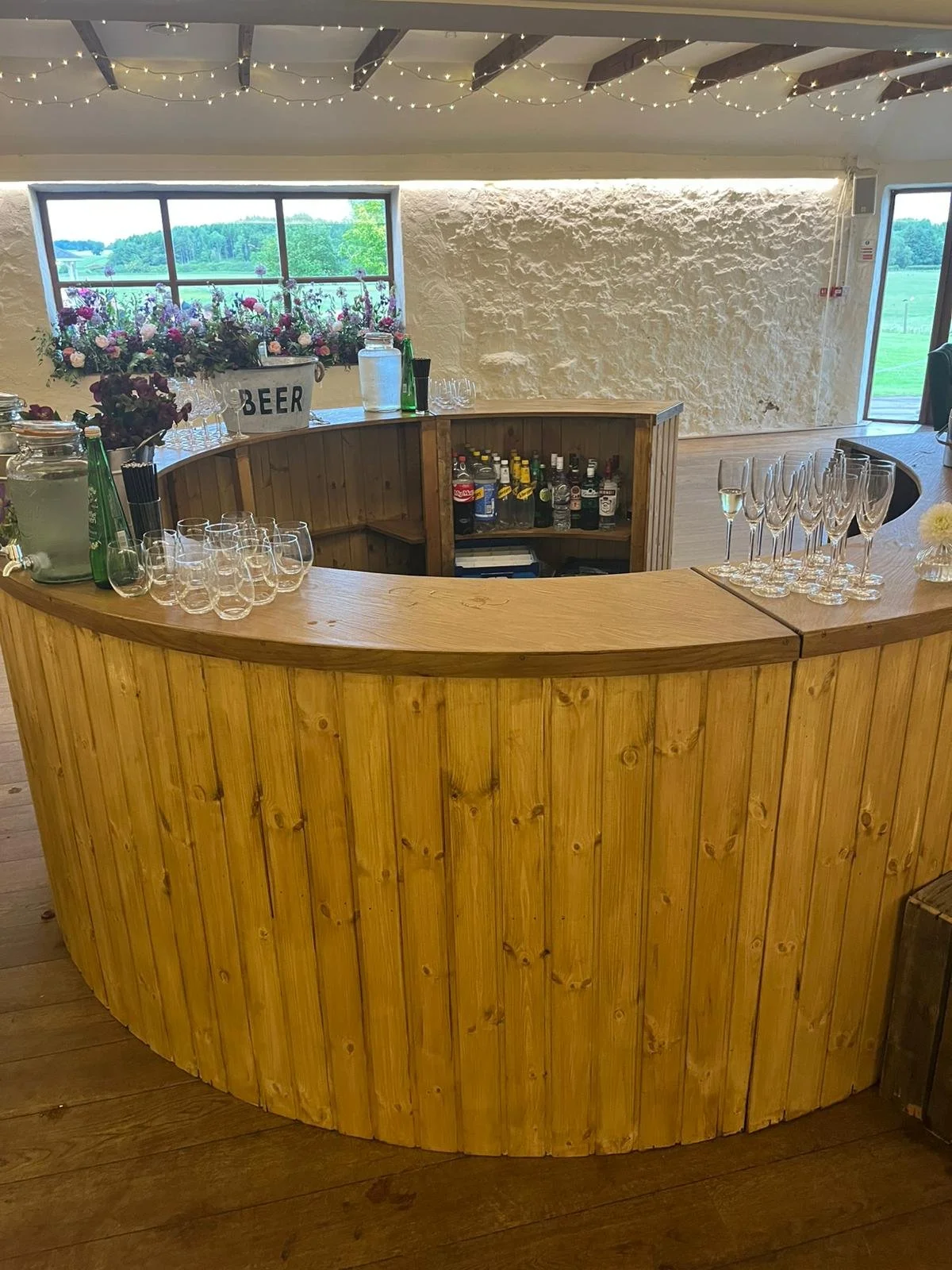 A wooden bar counter with glasses for drinks, bottles of liquor behind it, and flowers with a 'Beer' sign on the window ledge; fairy lights hanging from the ceiling and windows showing a green outdoor landscape.