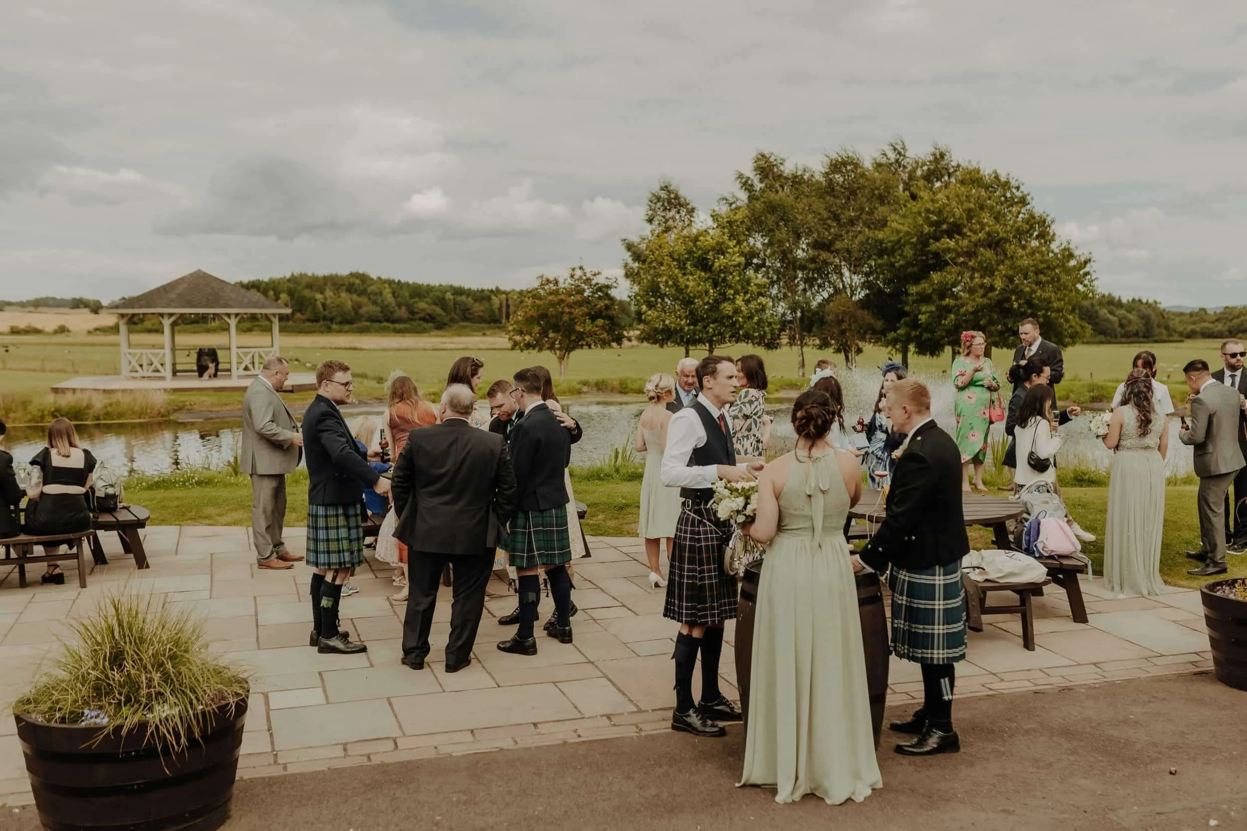Wedding reception outdoors by a pond with guests mingling, some in traditional Scottish kilts, in front of a scenic landscape with trees and a gazebo.