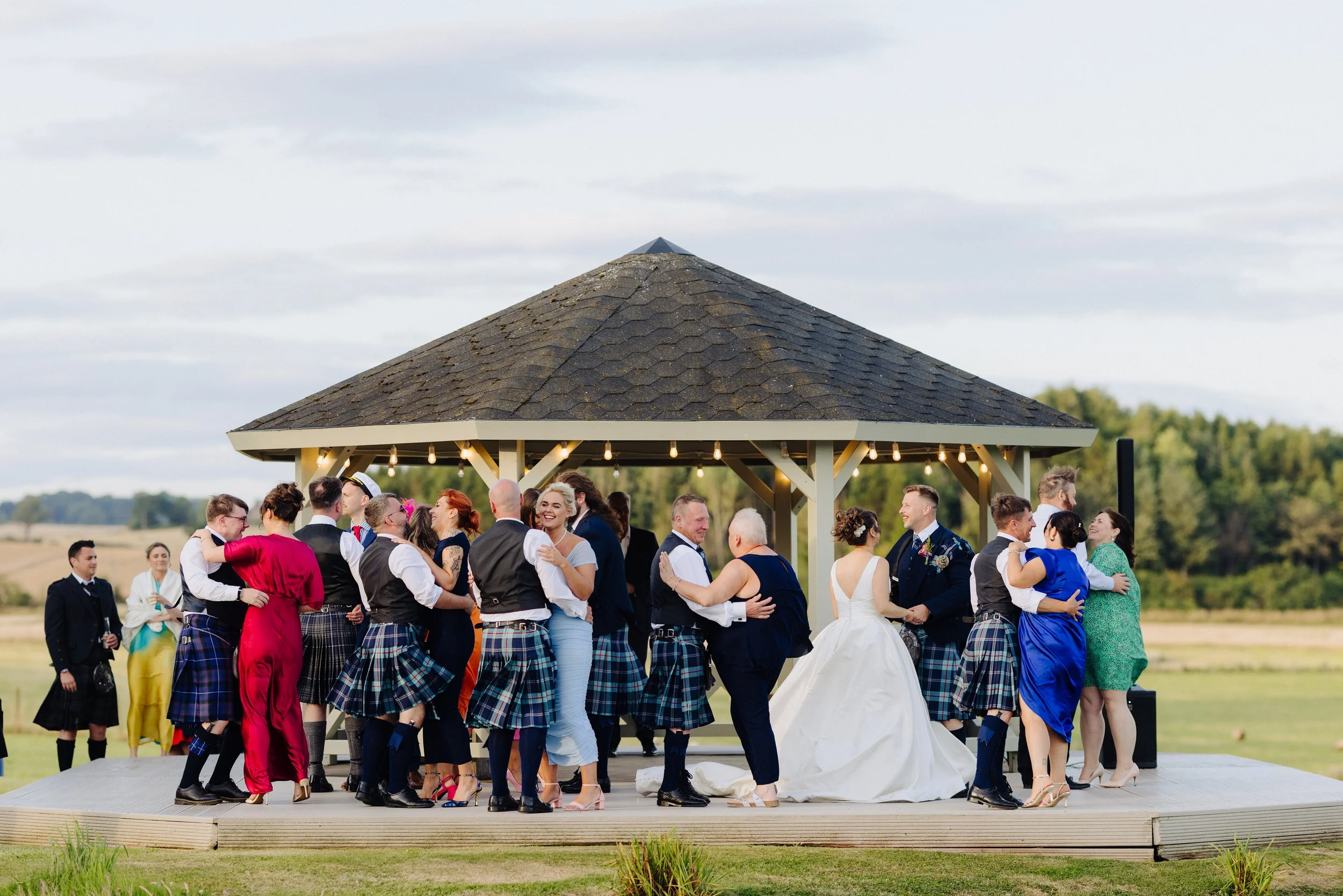 People dancing at an outdoor wedding reception under a pavilion with string lights, surrounded by green fields and trees.