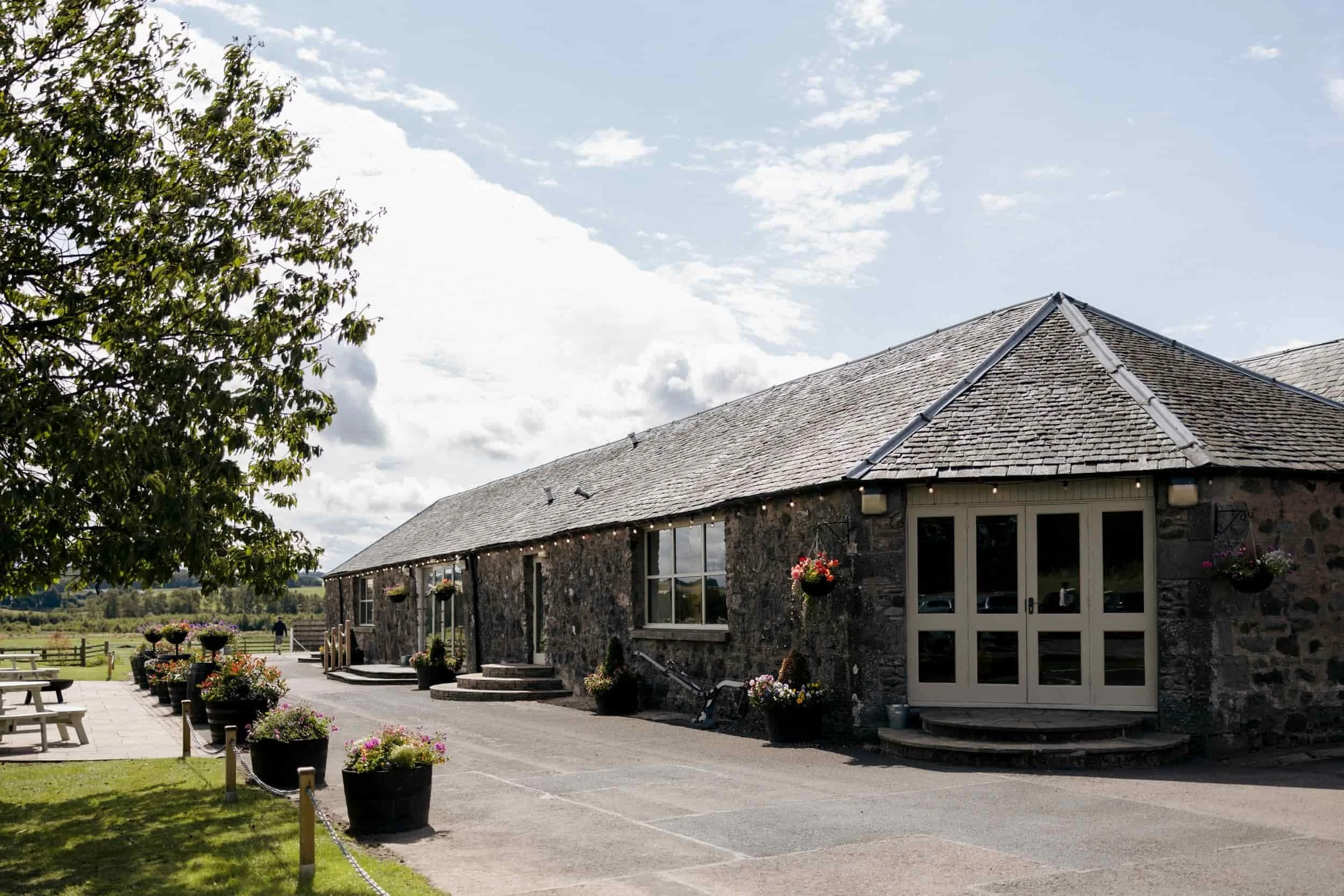 Stone building with a shingled roof, large glass doors, and flower baskets. Outdoor seating with benches, potted flowers, and a scenic view of a grassy field under a partly cloudy sky.