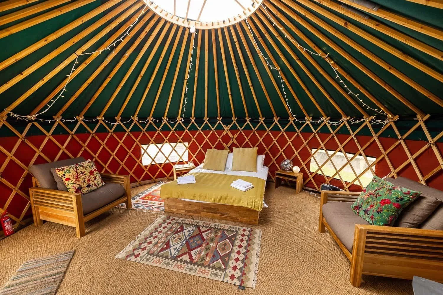 Interior of a yurt with a bed in the center, flanked by two couches with floral pillows, colorful rugs, and string lights hanging inside.