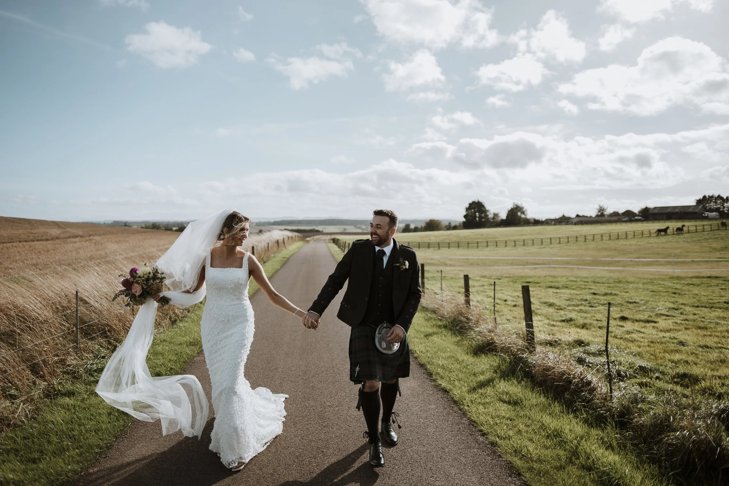 A bride and groom walking hand in hand on a rural road, smiling and holding a bouquet of flowers, under a partly cloudy sky.