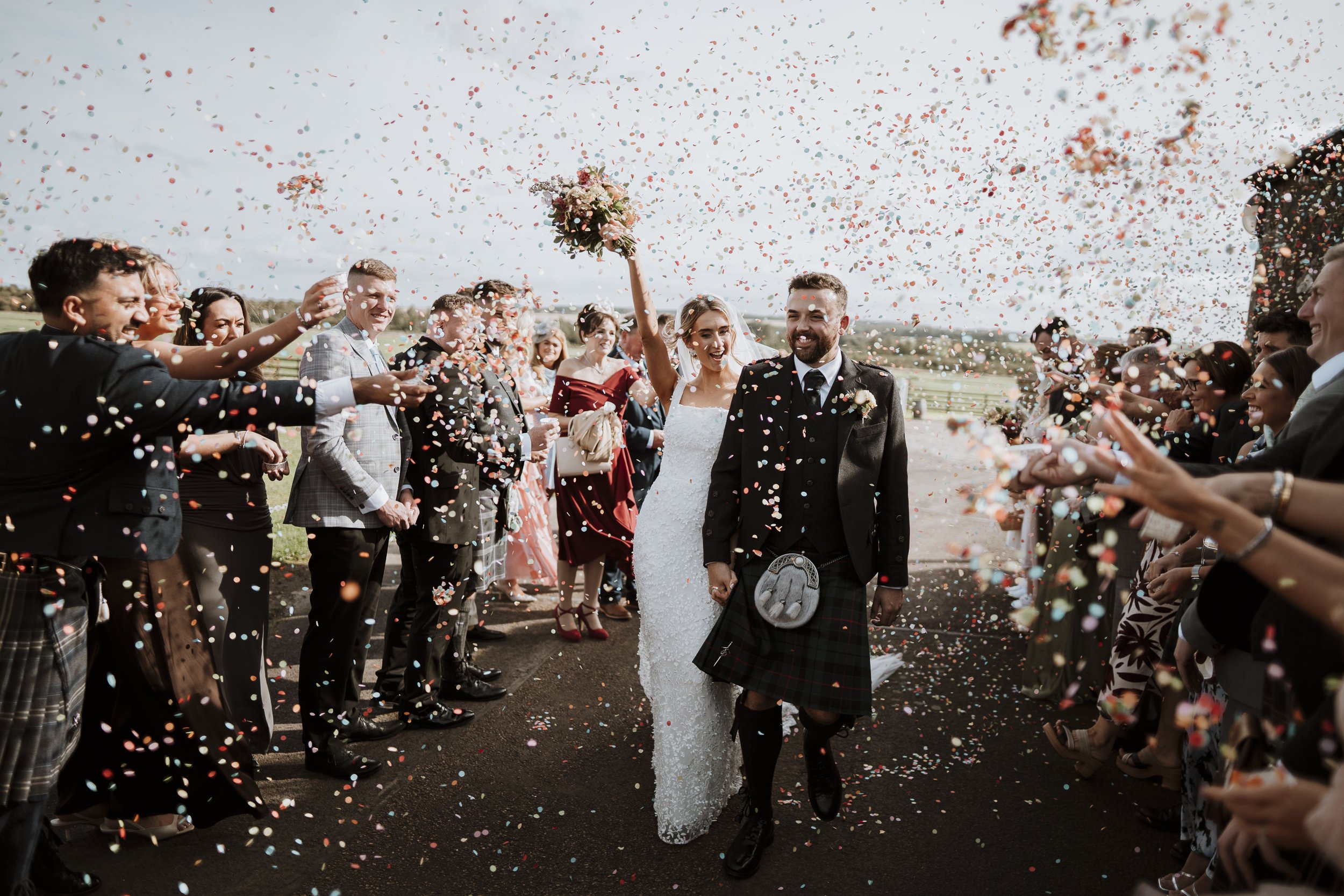 A bride and groom walking together under falling confetti, surrounded by celebrating guests at an outdoor wedding.
