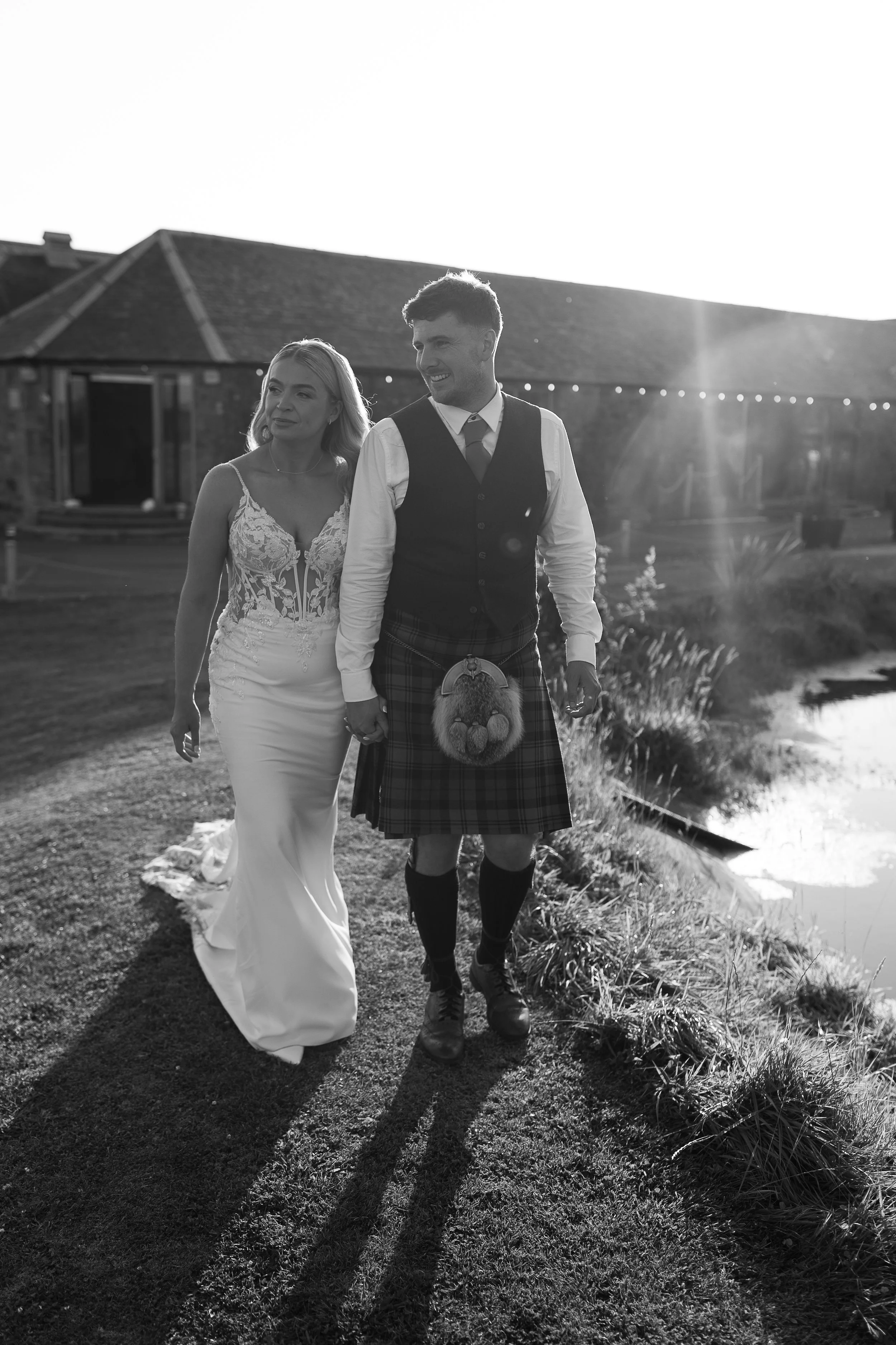 Black and white photo of a bride and groom walking outside during sunset, holding hands, with a building and a pond in the background.