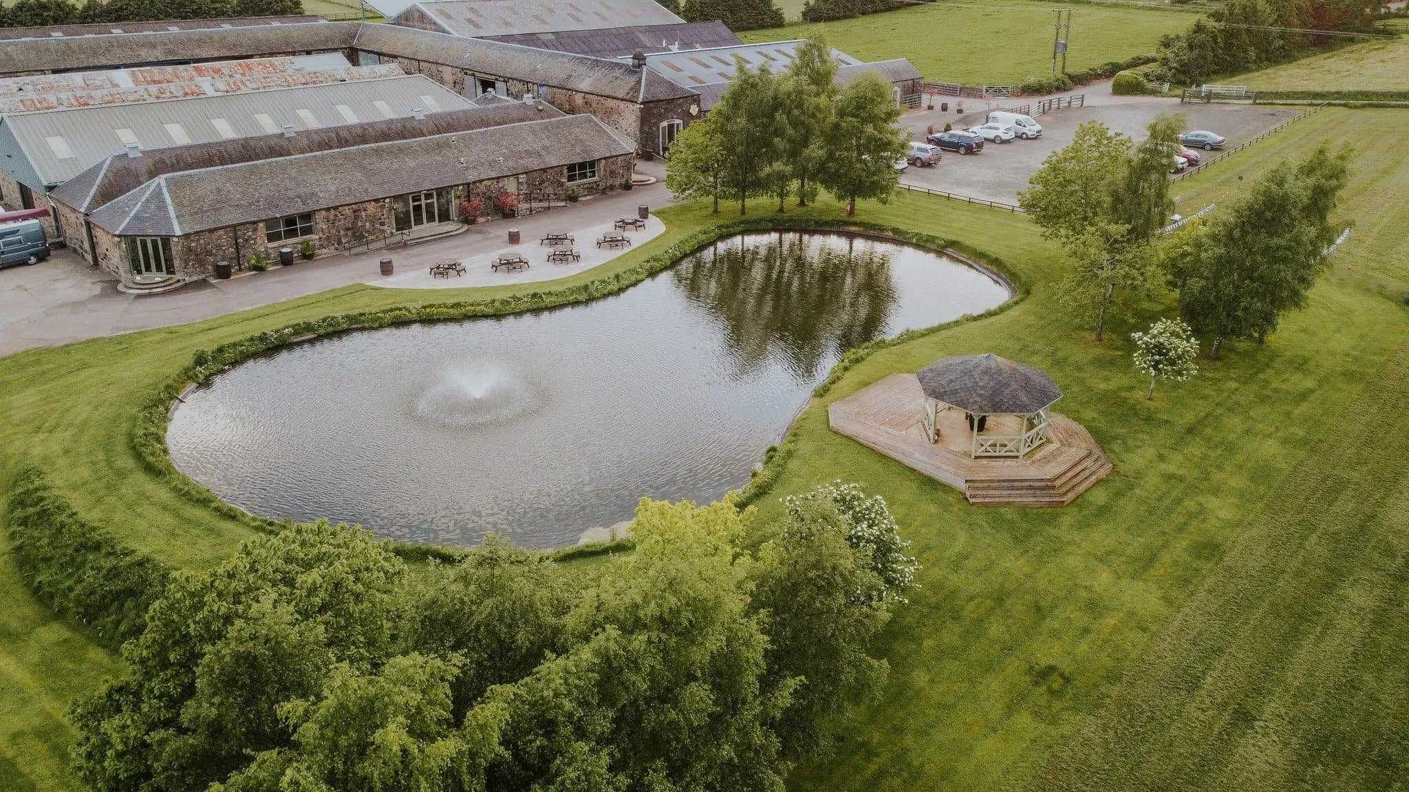 Aerial view of a pond with a fountain, a gazebo, and a stone building surrounded by green lawns and trees.