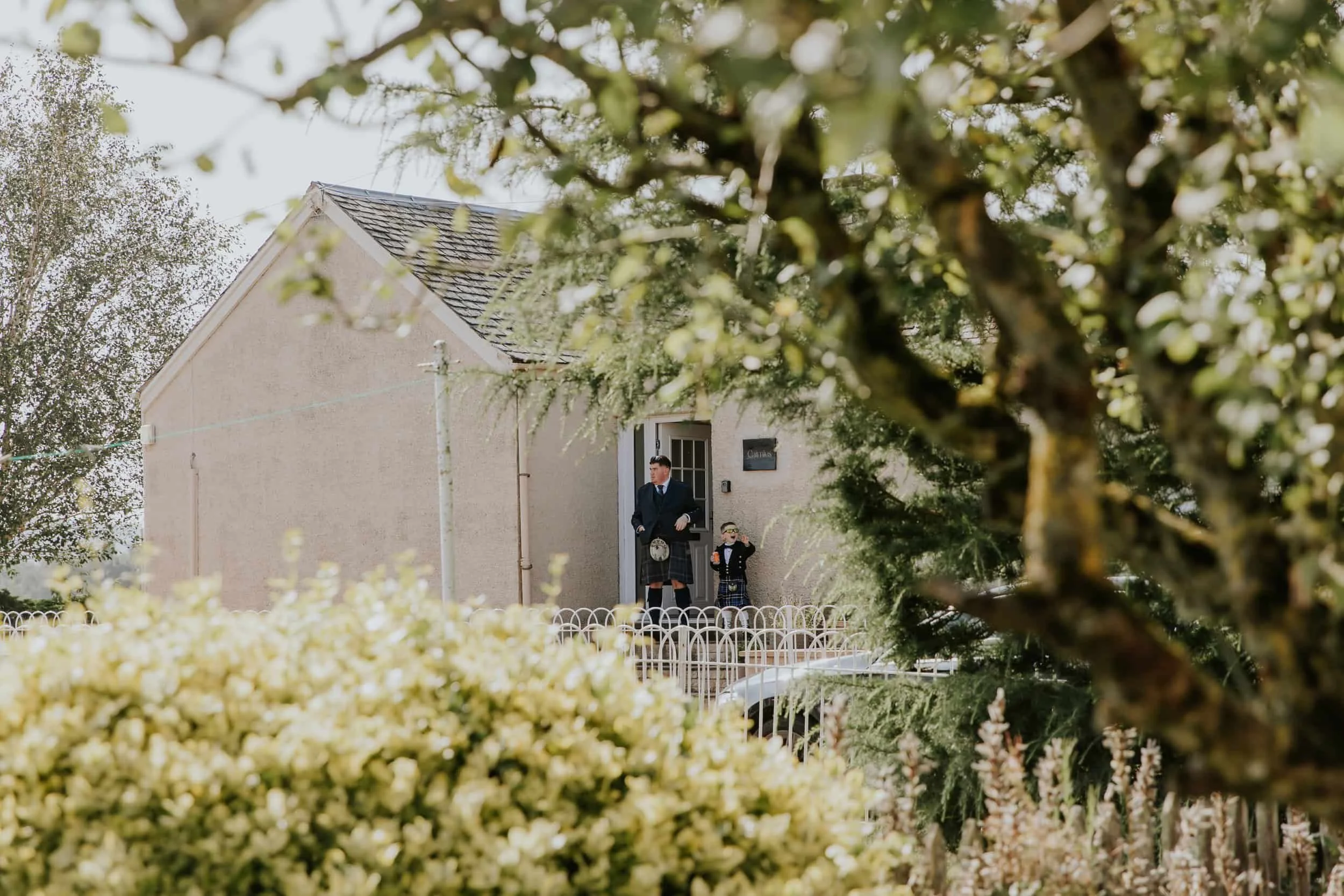 A man and a young boy dressed in traditional Scottish attire standing on the porch of a house, partially obscured by trees and bushes.