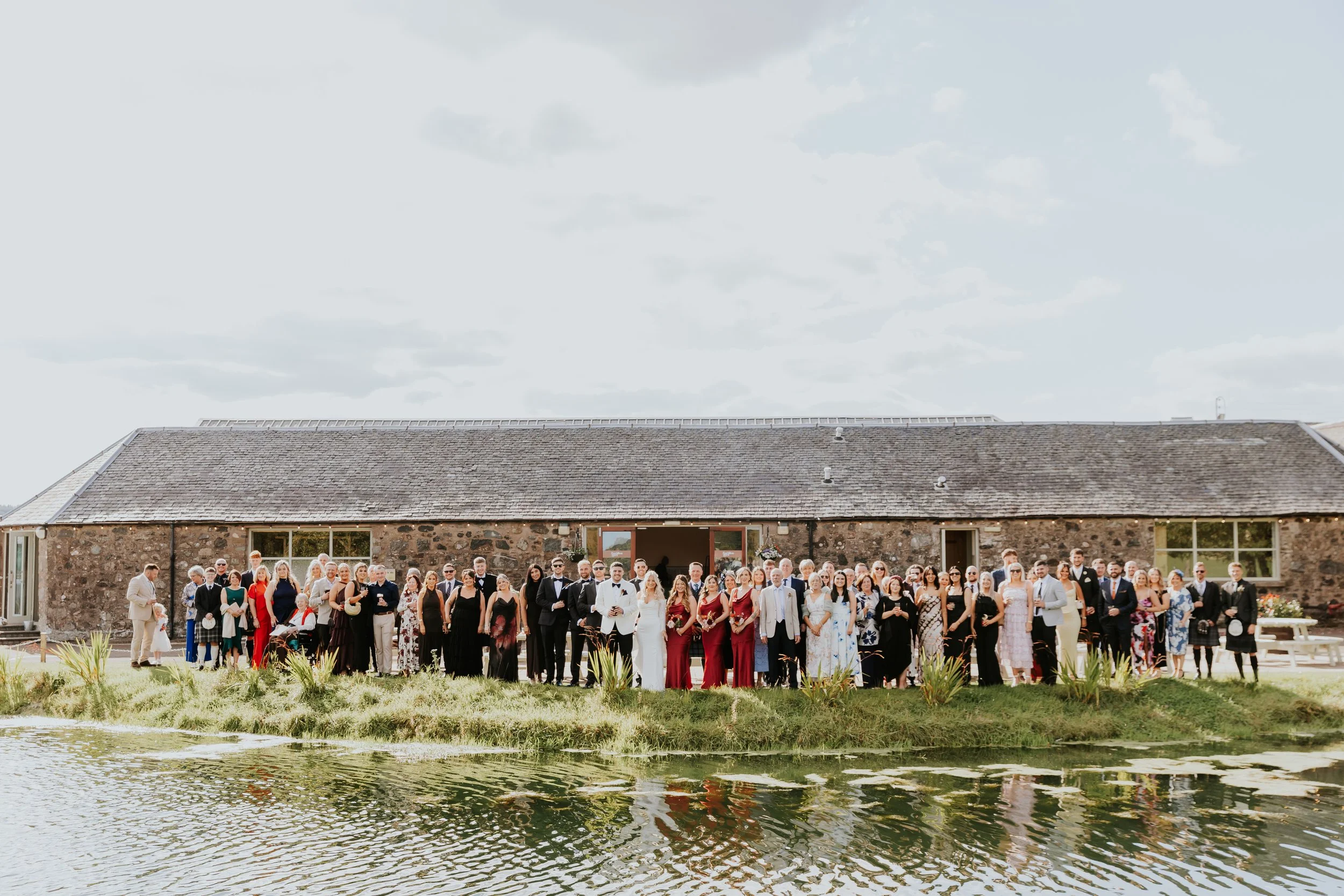 A large group of people dressed in formal attire standing in front of a rustic stone building by a pond on a cloudy day.