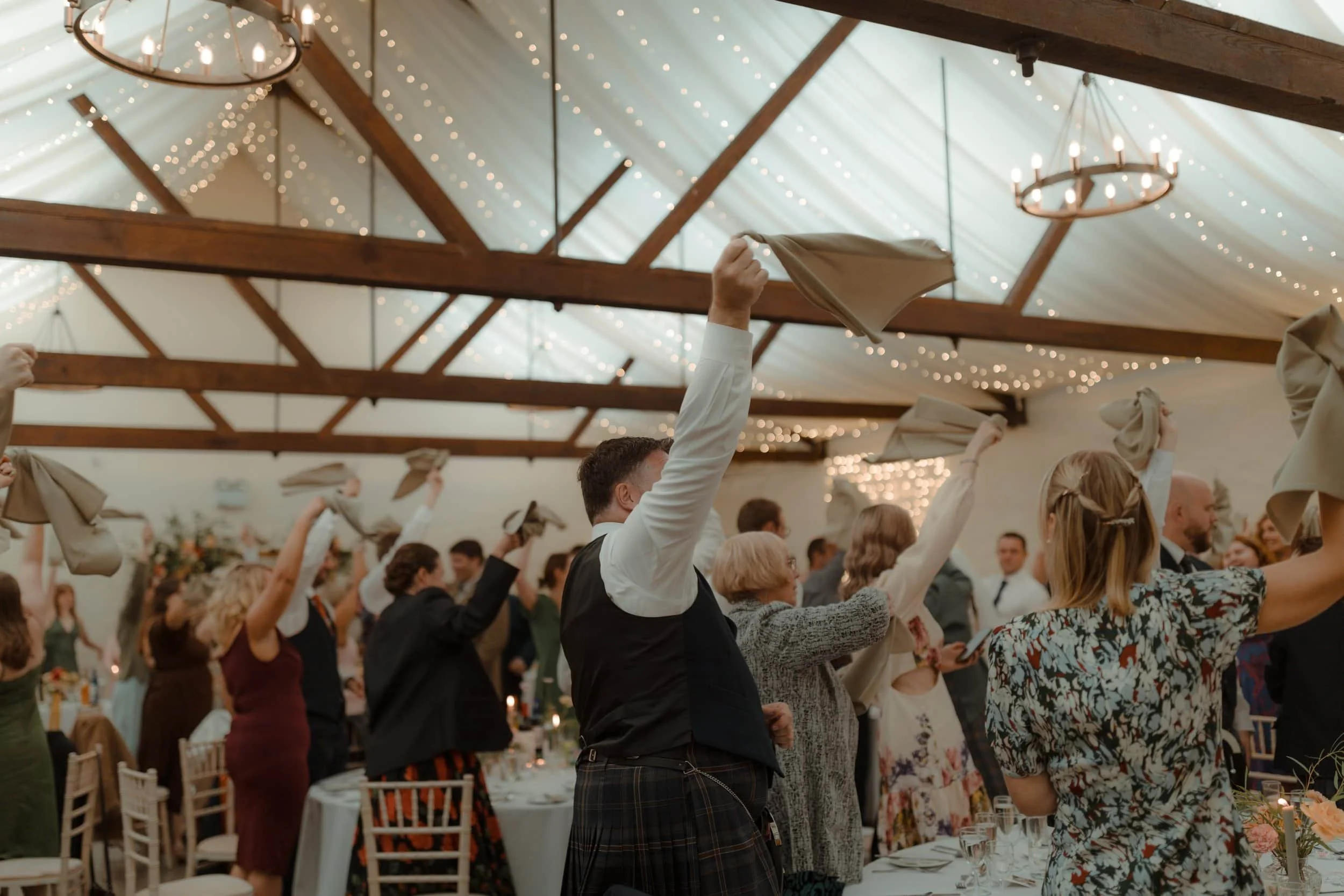 People dancing and raising napkins at a wedding reception or celebration in a decorated hall with string lights and wooden beams.