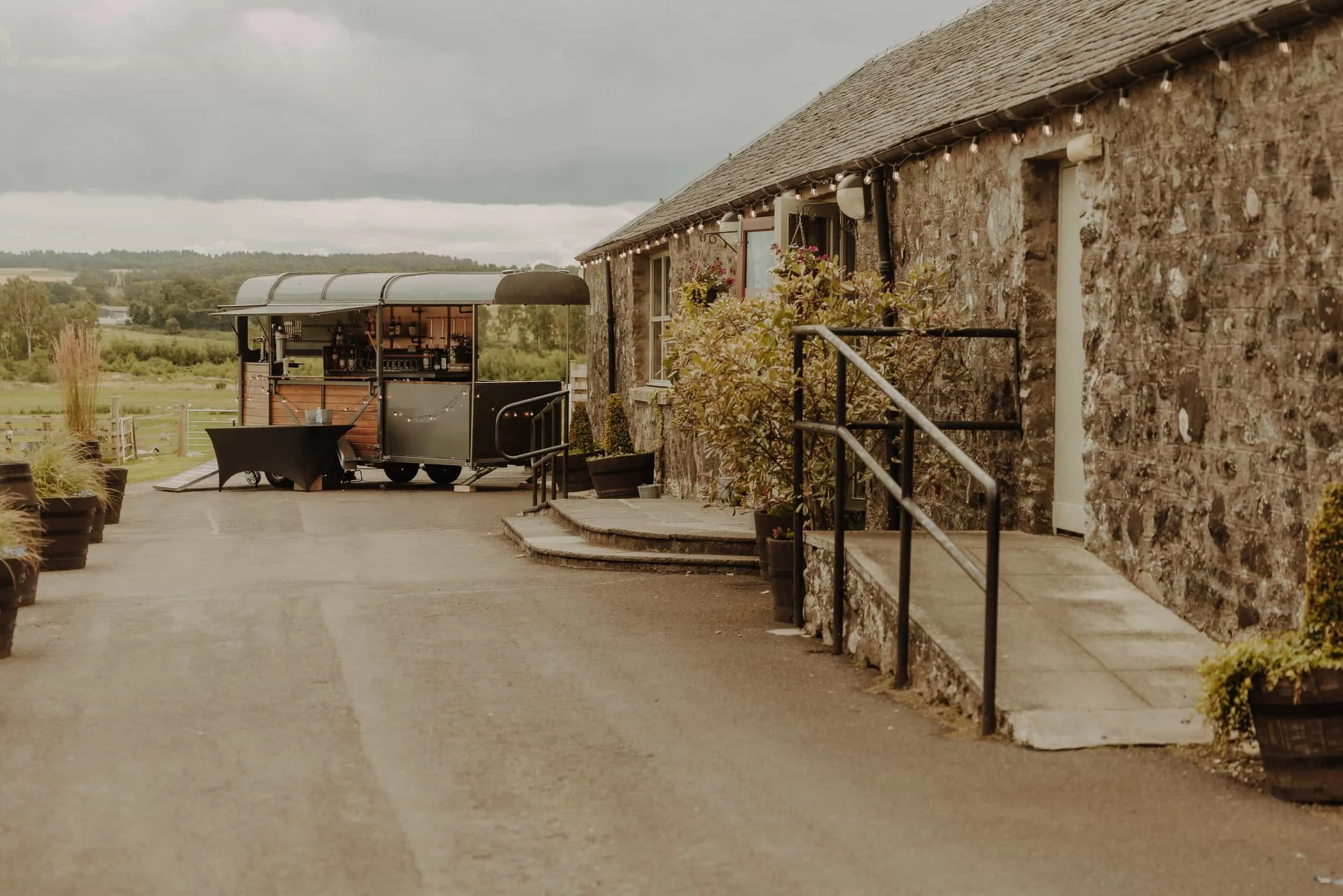 Outdoor patio area with a stone building and a food truck serving drinks, with potted plants and a scenic rural background under a cloudy sky.