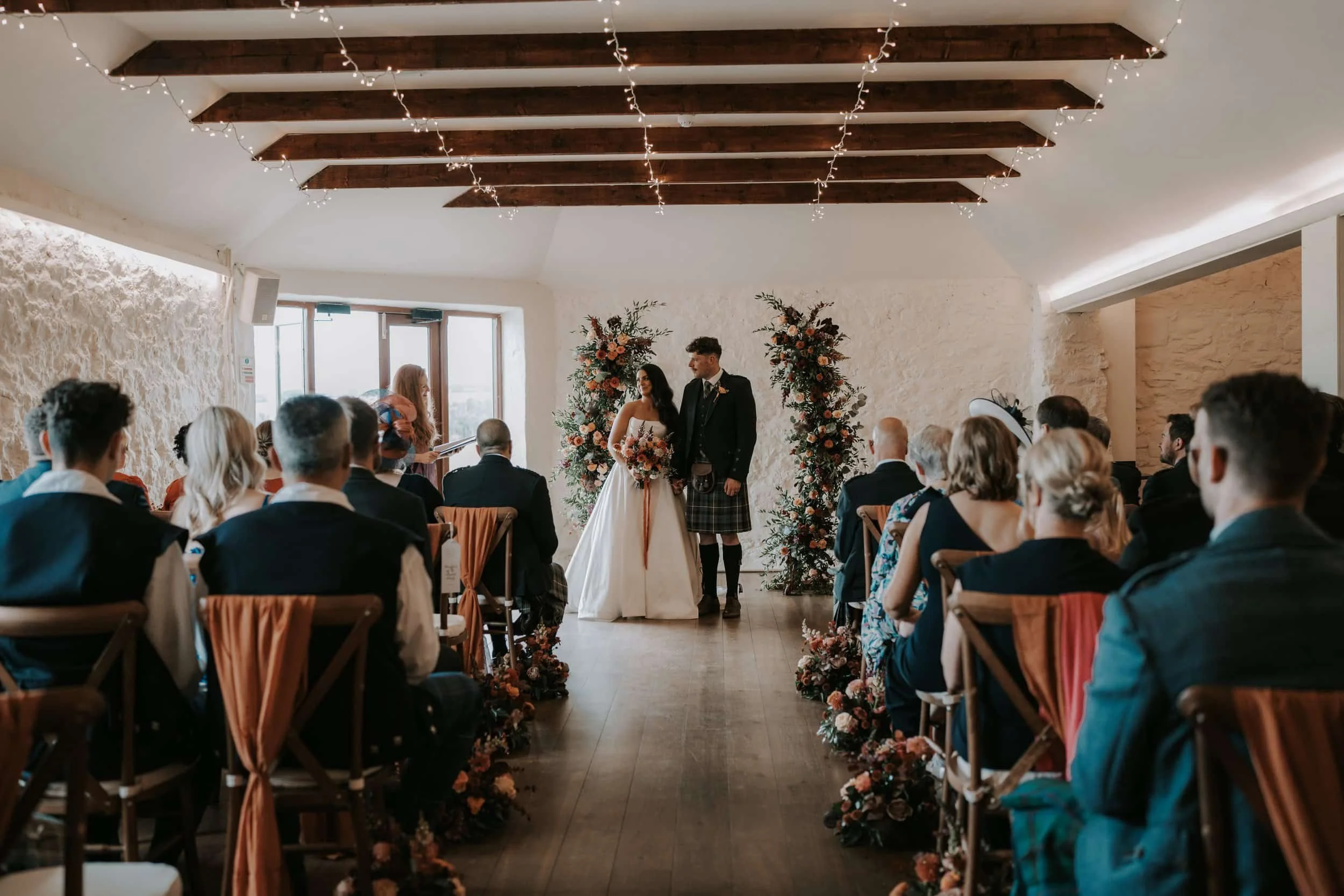 Couple exchanging vows during a wedding ceremony in front of floral arrangements, facing an officiant, with guests seated on either side in a cozy, rustic indoor space with string lights and wooden beams.