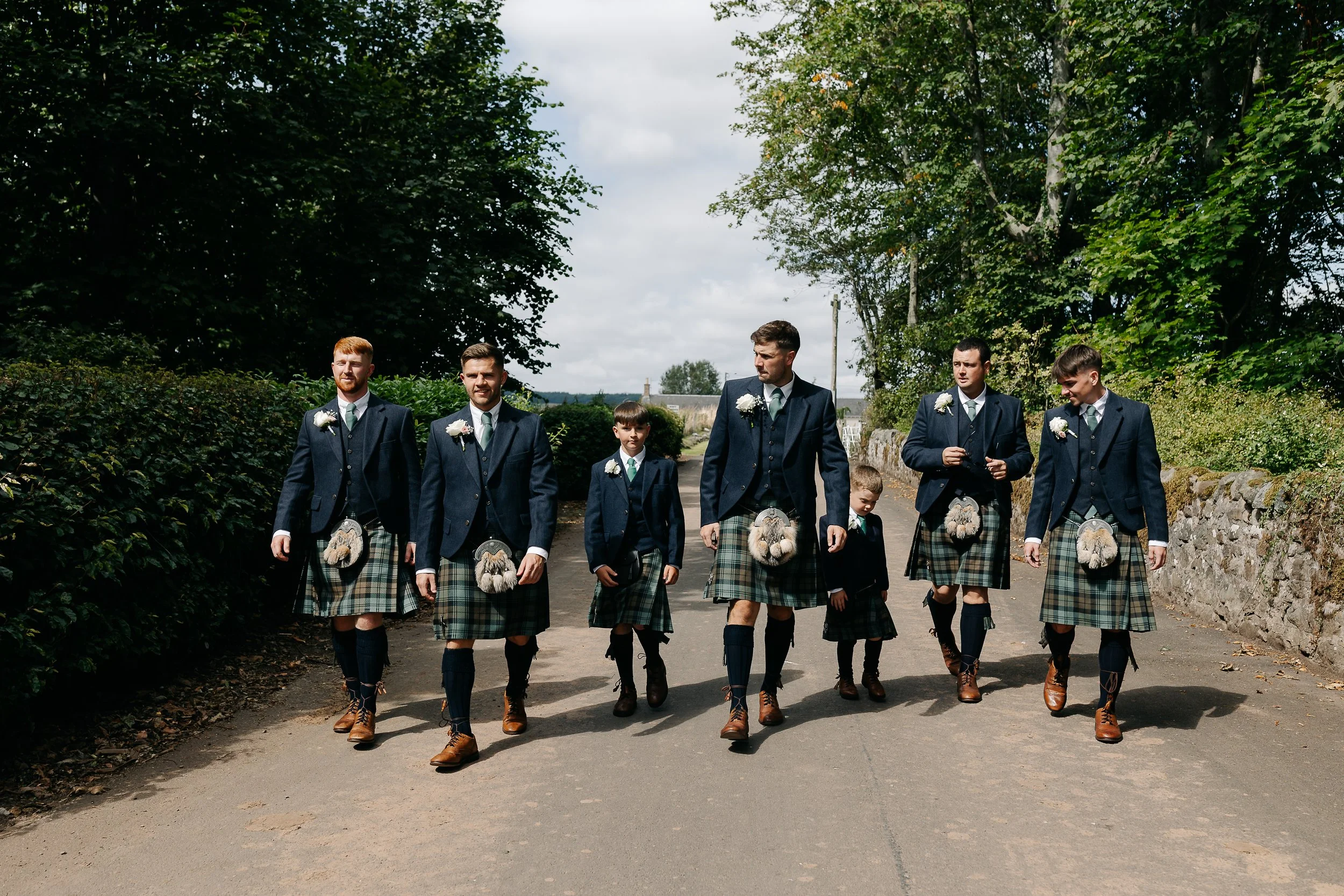 Men and boys wearing traditional Scottish kilts and jackets walking on a rural road.