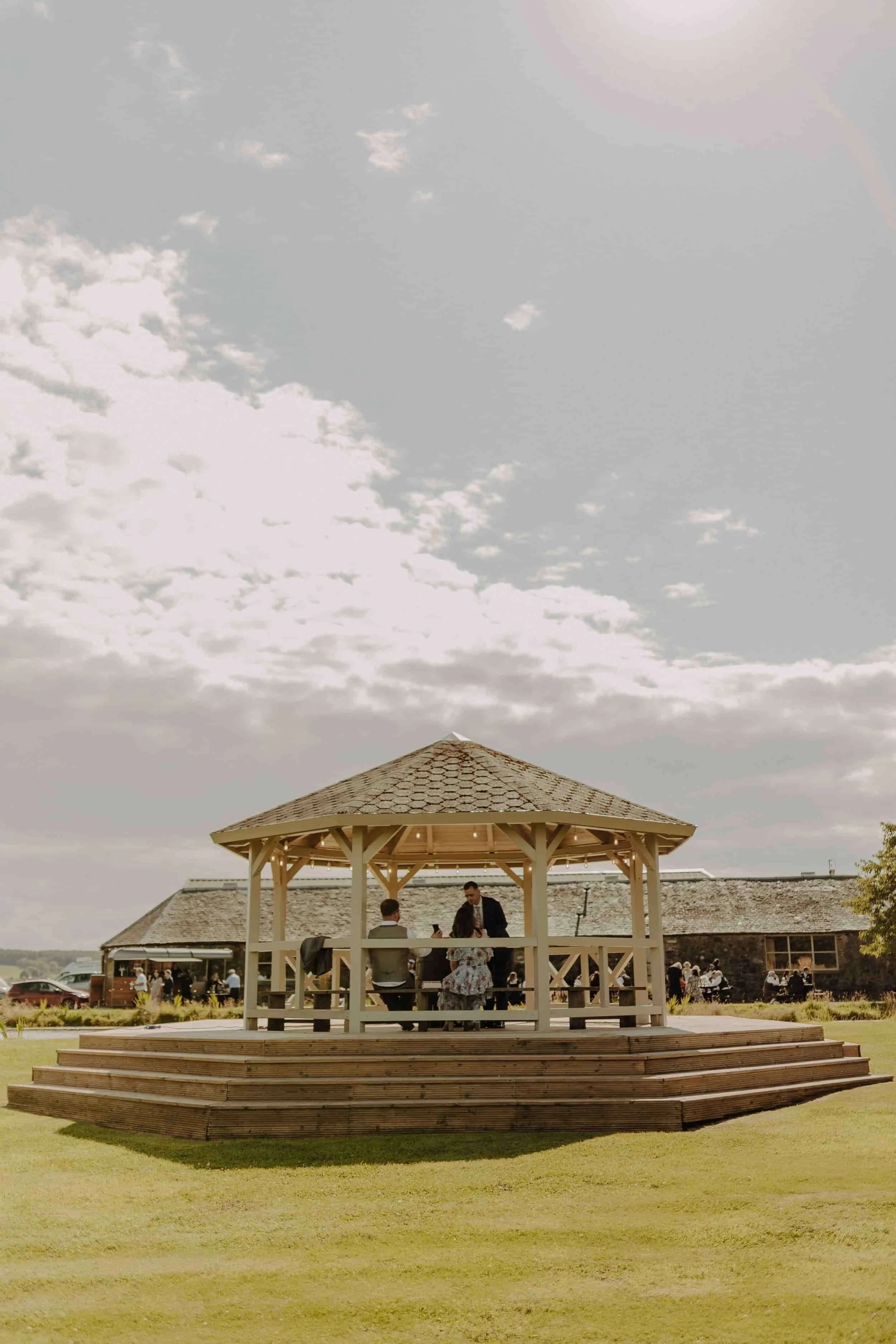 People gathered under a wooden gazebo outdoors during the daytime, with a building and parked cars in the background.