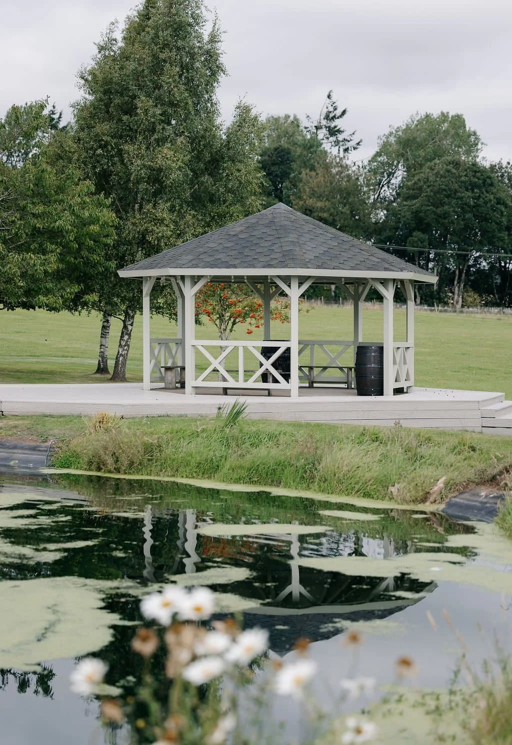 A white gazebo with a gray shingled roof is situated on a grassy area next to a pond with lily pads and white flowers. There are trees in the background under a cloudy sky.