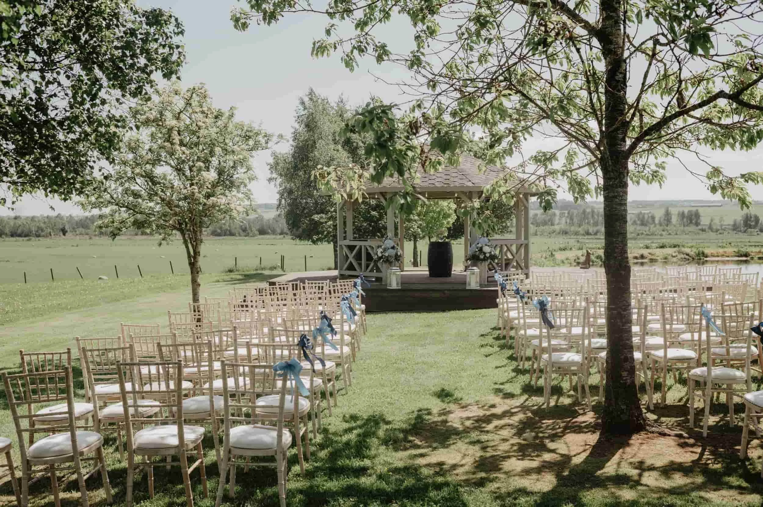 Outdoor wedding ceremony setup with white chairs decorated with blue ribbons, facing a gazebo on a grassy field, surrounded by trees and scenic countryside under a clear sky.