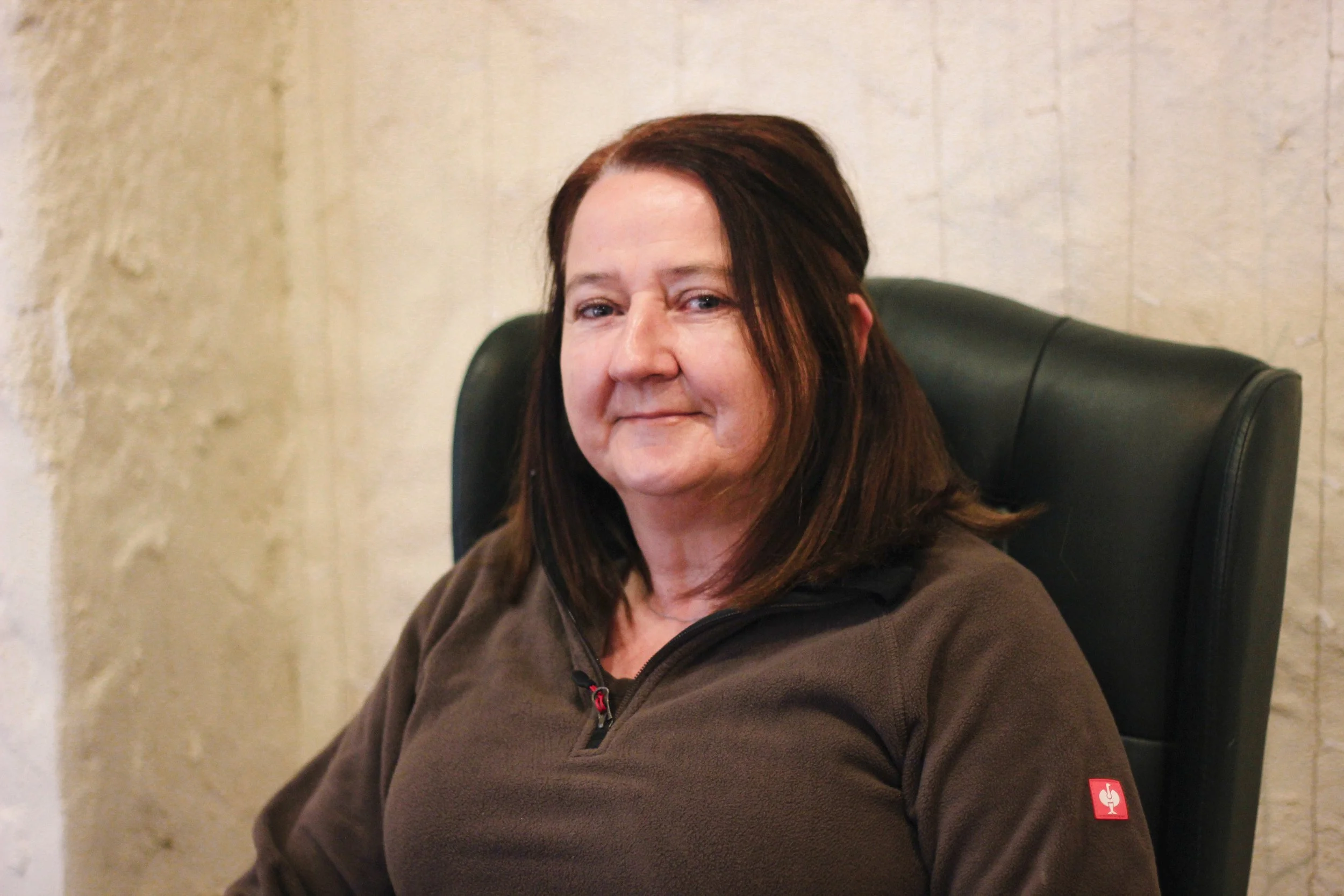 A woman with shoulder-length brown hair sitting in a black office chair, wearing a brown fleece jacket with a red logo on the sleeve, smiling slightly, in an indoor setting with a beige textured wall in the background.