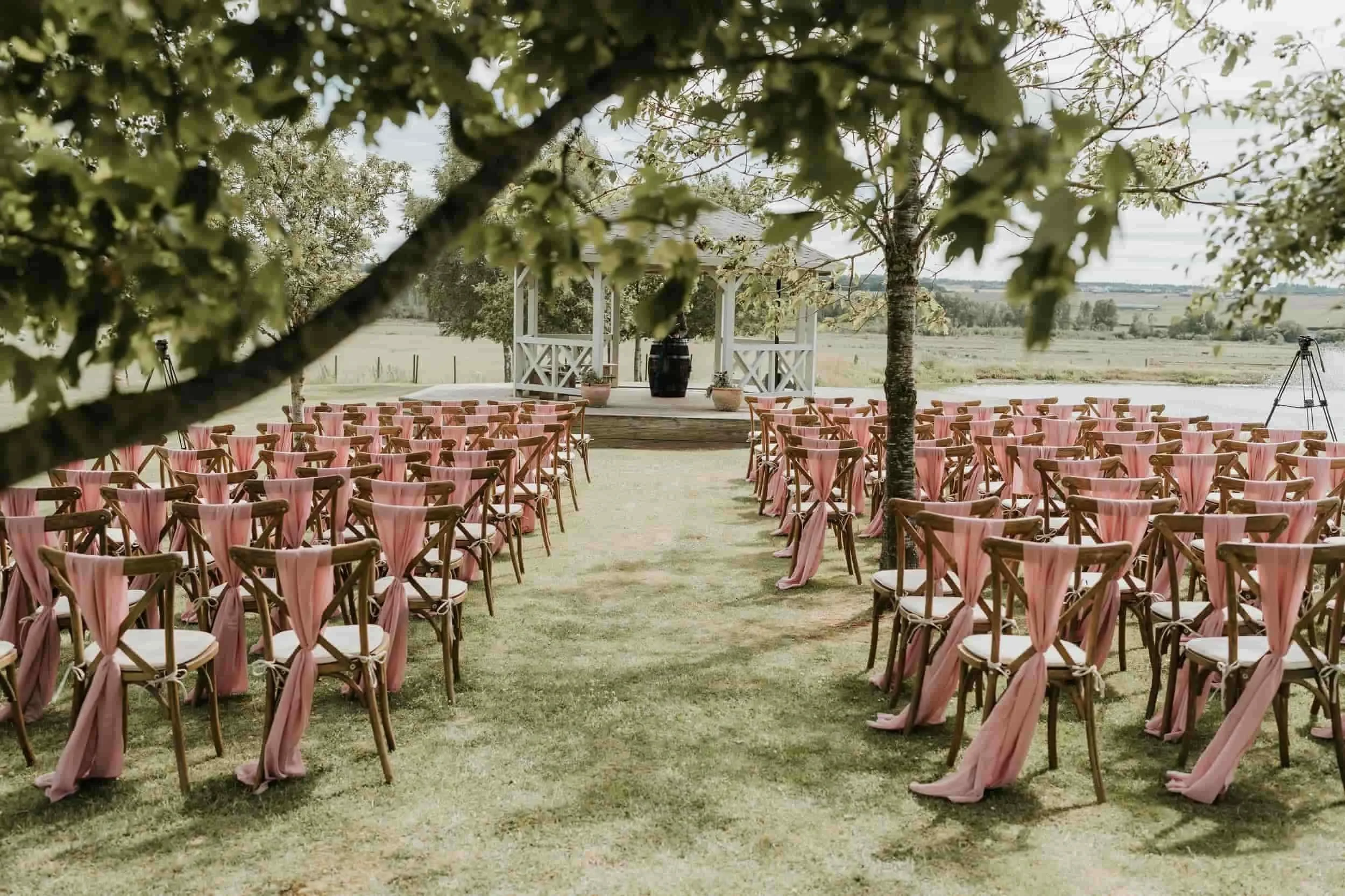Outdoor wedding ceremony setup with wooden chairs decorated with pink fabric, arranged on a grassy area facing a small white pavilion with a lake and trees in the background.