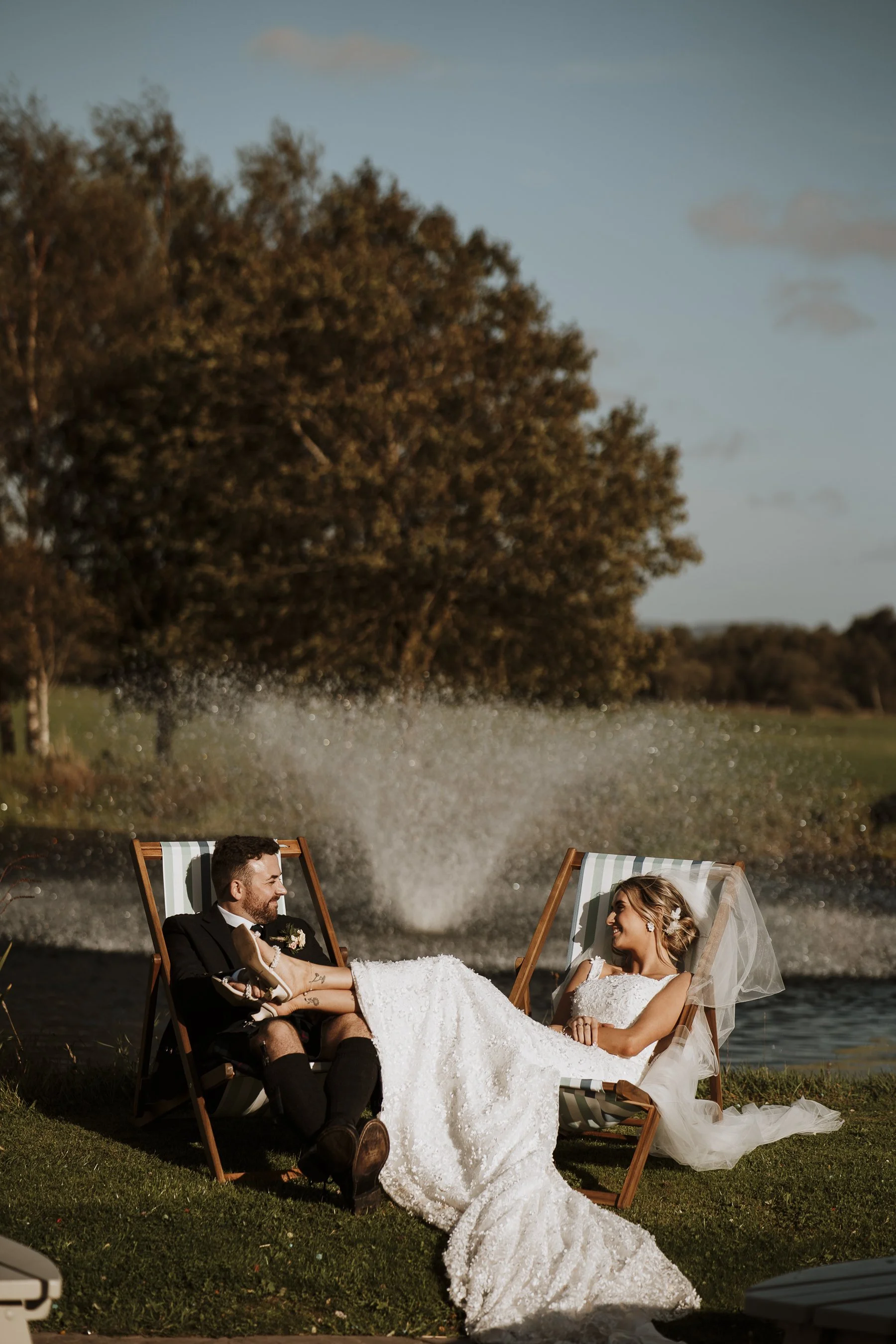 A bride and groom sitting together on lounge chairs near a lake, with a boat splash behind them; the bride is wearing a veil and white wedding dress, and the groom is in a suit with shorts, both smiling and enjoying the moment.