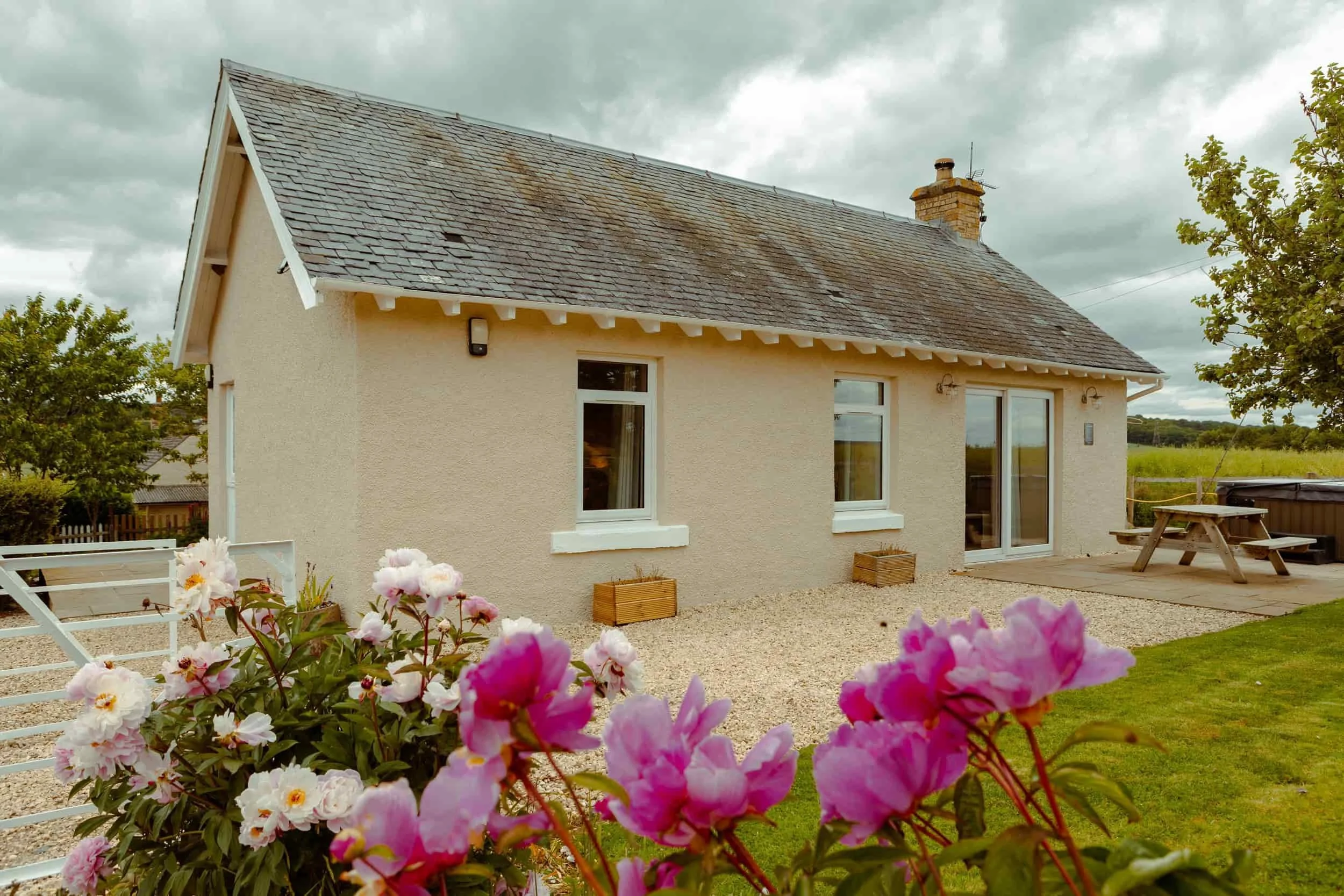 Small beige cottage with white trim, surrounded by flowers and greenery under a cloudy sky, with a picnic table outside.