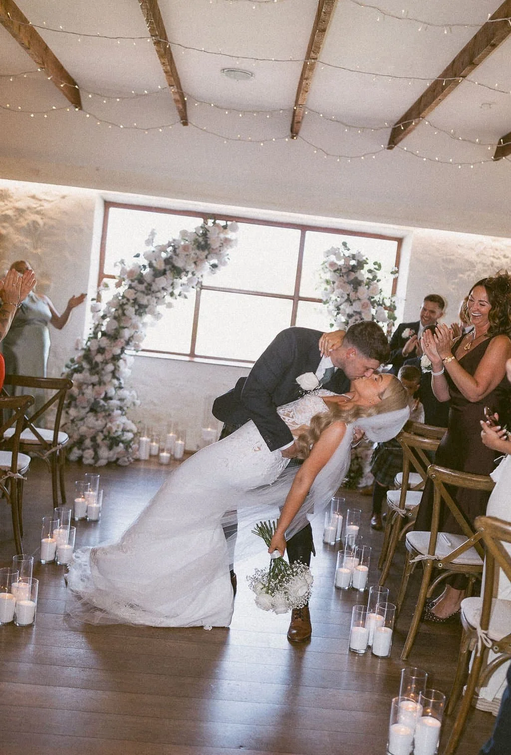A newlywed couple sharing a dip kiss during their wedding reception, surrounded by friends and family in a decorated indoor space with candles and floral arrangements.