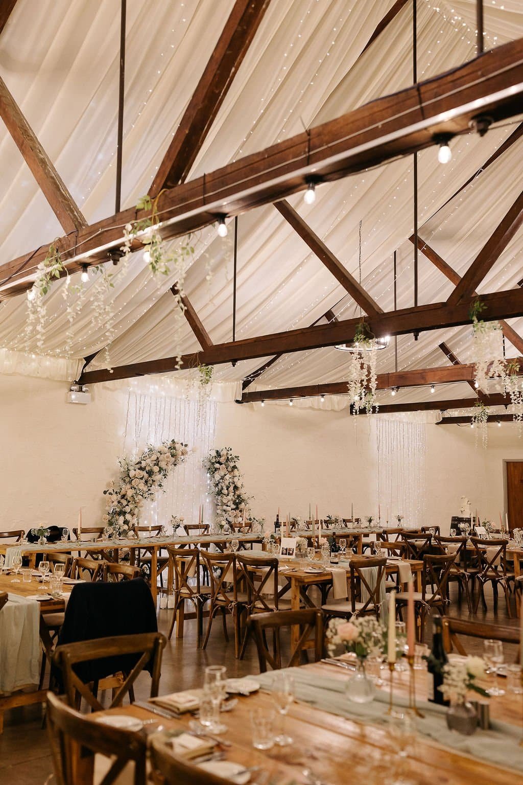 Elegant indoor wedding reception setup with long wooden tables, floral centerpieces, and string lights hanging from a wooden-beamed ceiling decorated with hanging white flowers and greenery.