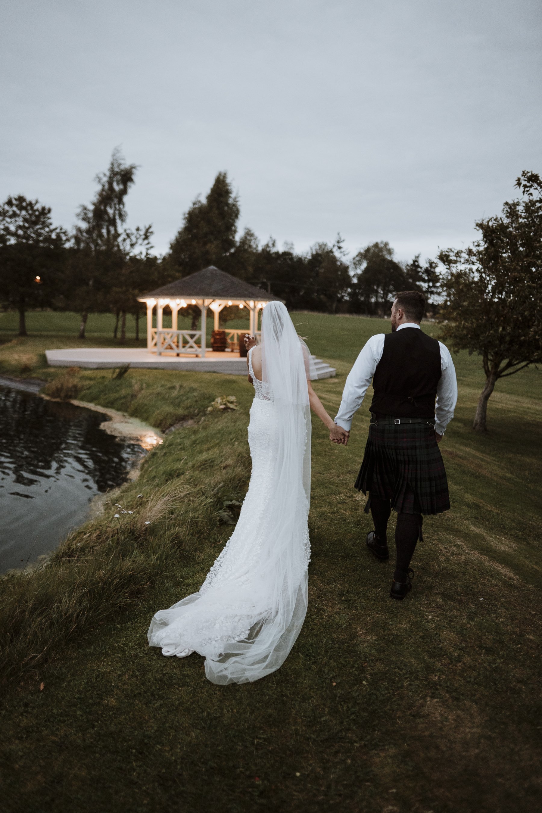 A bride and groom holding hands walking along a grassy path near a small pond at dusk with a lit gazebo in the background.